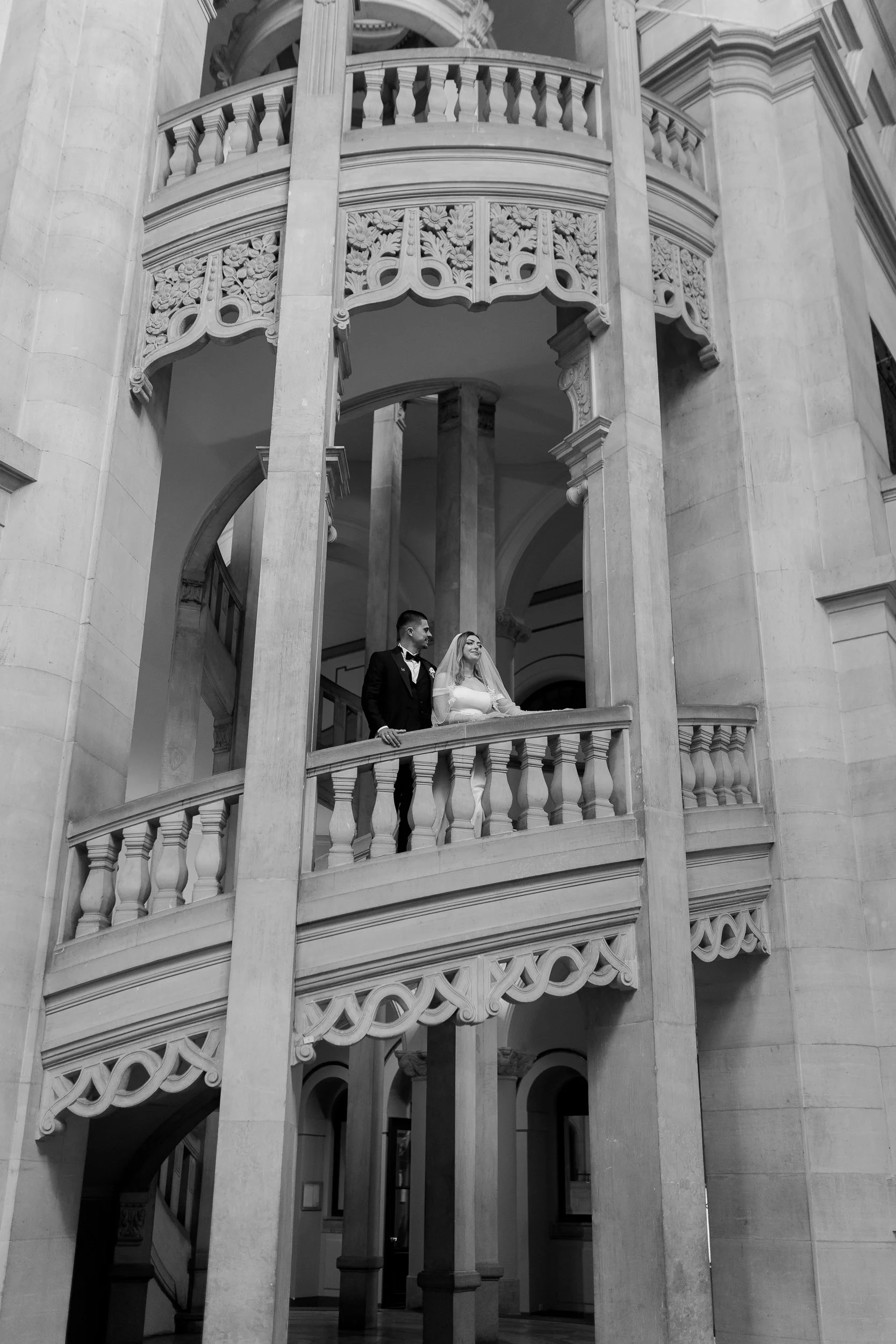 Zwei Menschen, ein Mann und eine Frau, auf einem Balkon in einem prächtigen, historischen Gebäude mit kunstvoller Architektur, vermutlich bei einer Hochzeit.