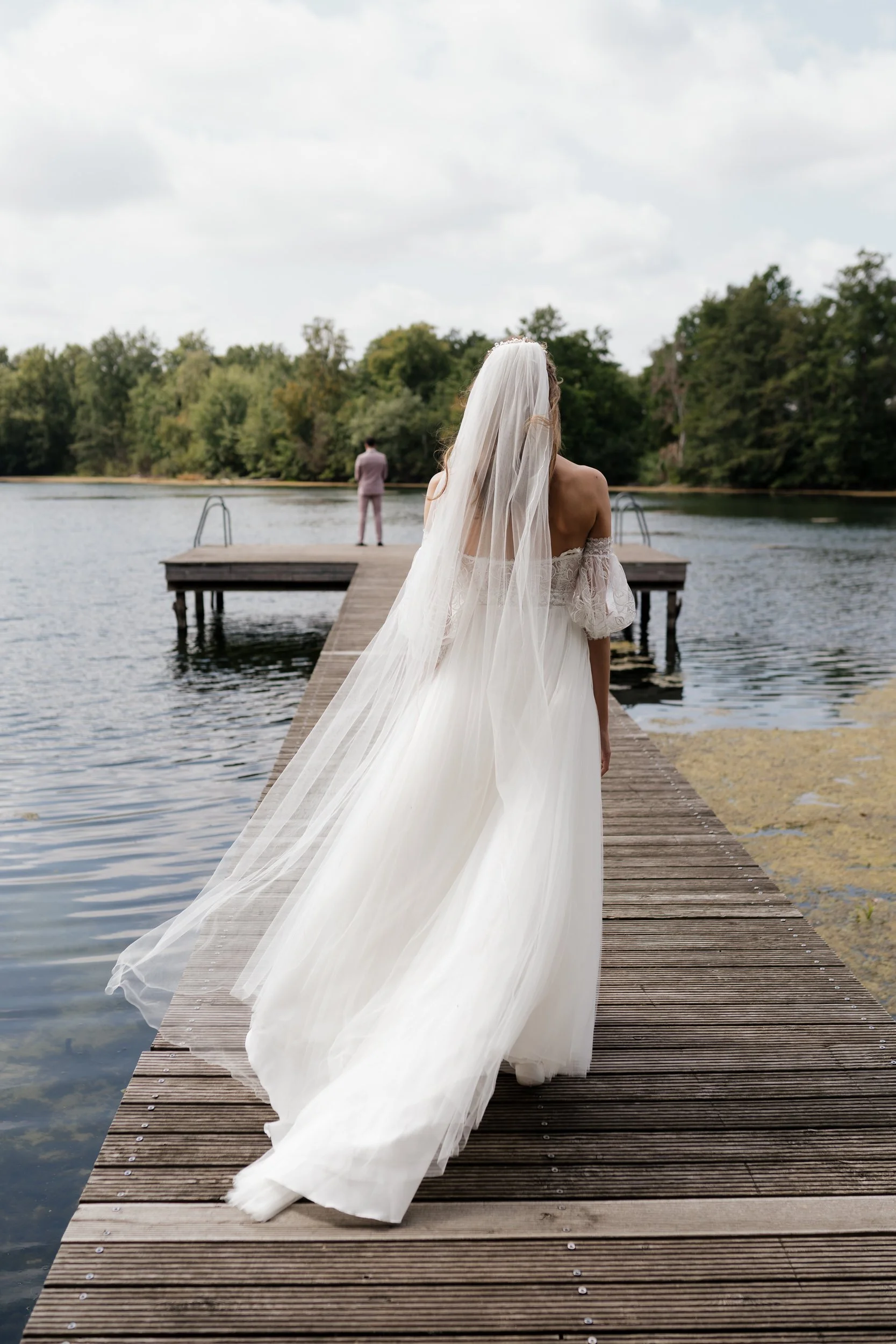 Braut in weißem Brautkleid mit langer Schleppe und Schleier steht auf einem Steg am Wasser, während der Bräutigam in Hellgrauem Anzug am Ende des Stegs steht und in die Ferne blickt.