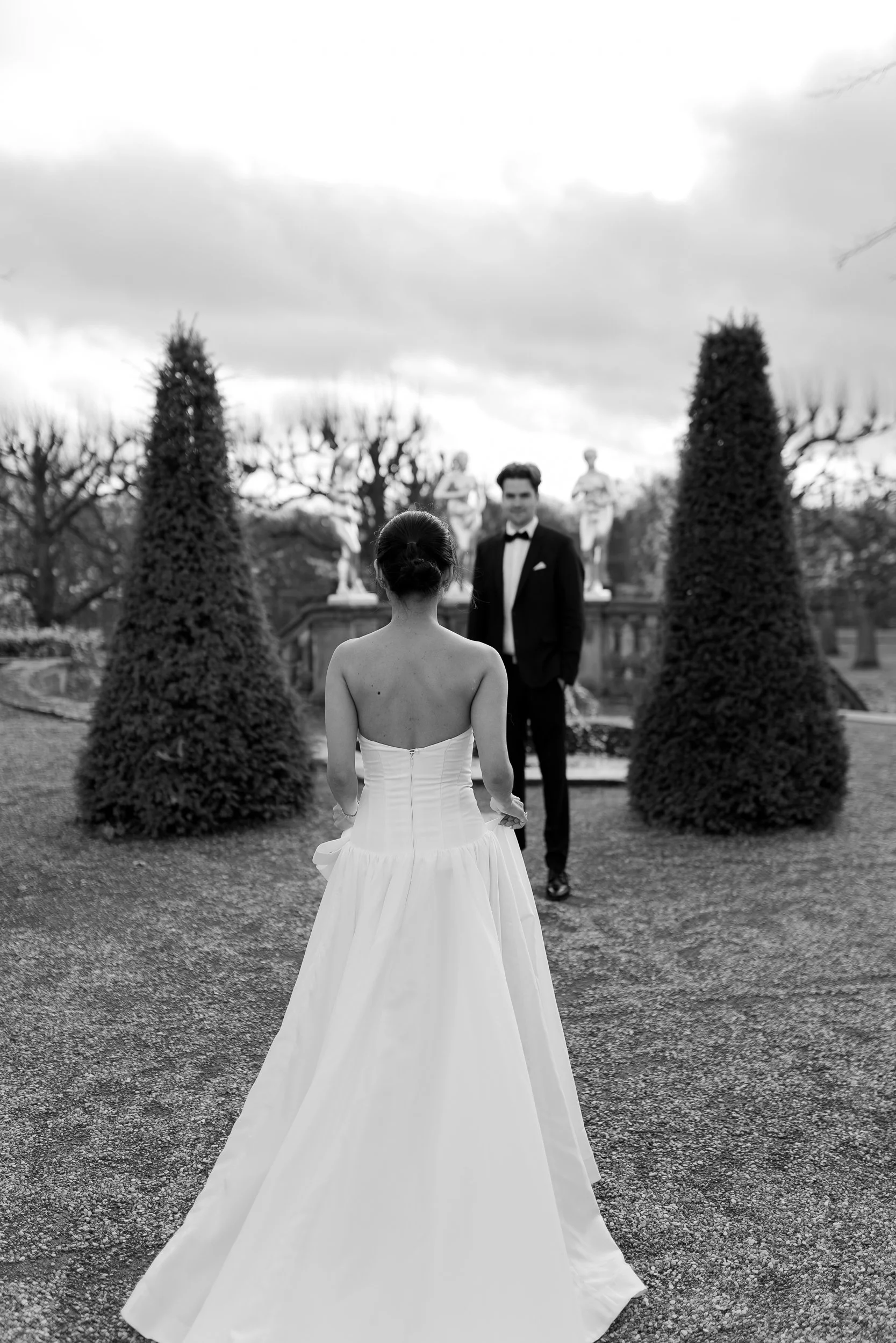 Braut mit Hochzeitskleid steht vor einem Mann in Anzug in einem Garten mit Sträuchern und Statuen, Himmel mit Wolken im Hintergrund, schwarz-weiß Bild.