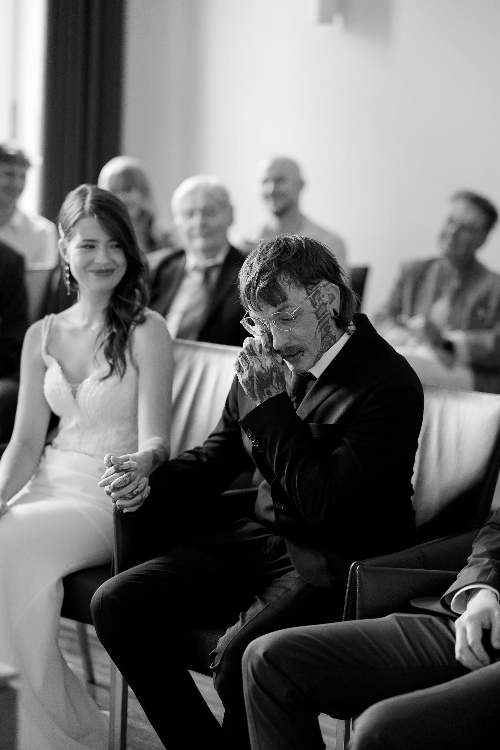 Schwarz-Weiß-Foto von einer Gruppe von Menschen, die bei einer Veranstaltung sitzen, darunter ein rosa-haariger Mann mit Tattoos, der den Kopf auf die Hand stützt, und eine lächelnde Frau in einem weißen Kleid.