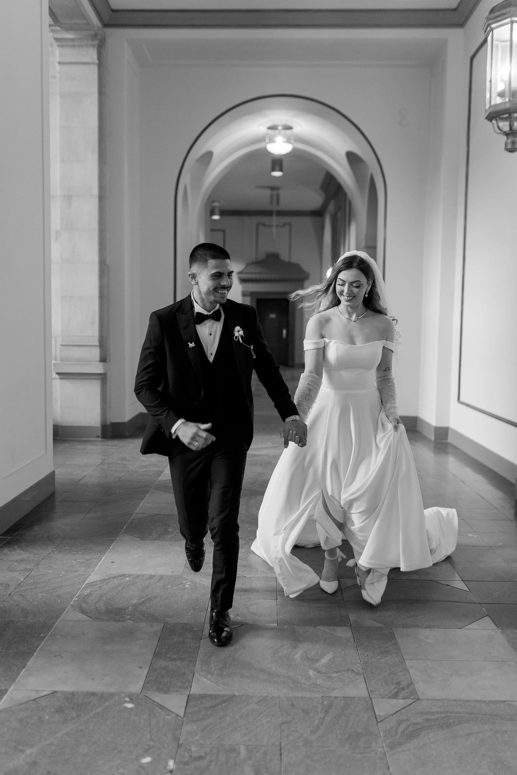 Ein glückliches Hochzeitspaar läuft Hand in Hand durch einen Flur in einem eleganten Gebäude, beide lächeln. Die Braut trägt ein langes weißes Kleid, der Bräutigam einen schwarzen Anzug mit Fliege.