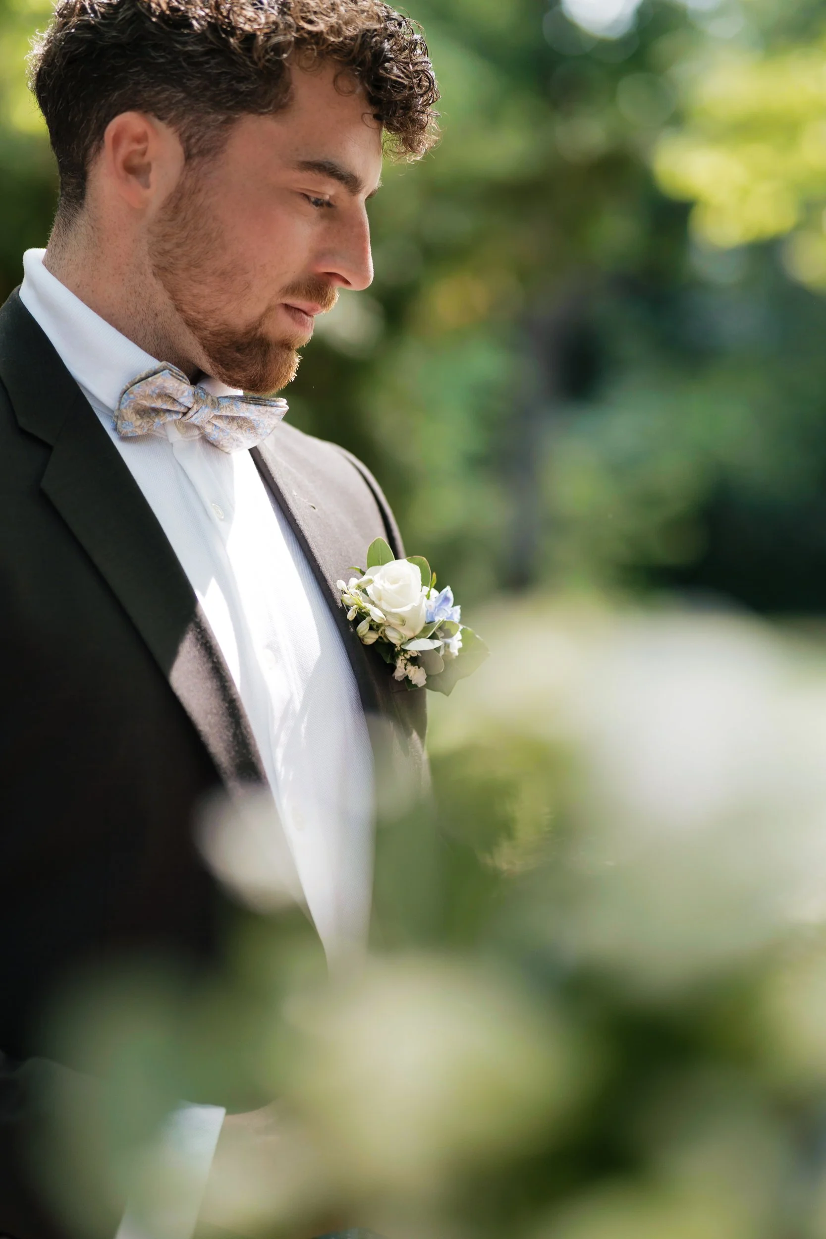 Ein junger Mann in Anzug, mit einer bunten Fliege, steht im Freien mit einem weißen Blumenboutonnière an der Brust, umgeben von grünen Bäumen, bei einer Hochzeitsveranstaltung.