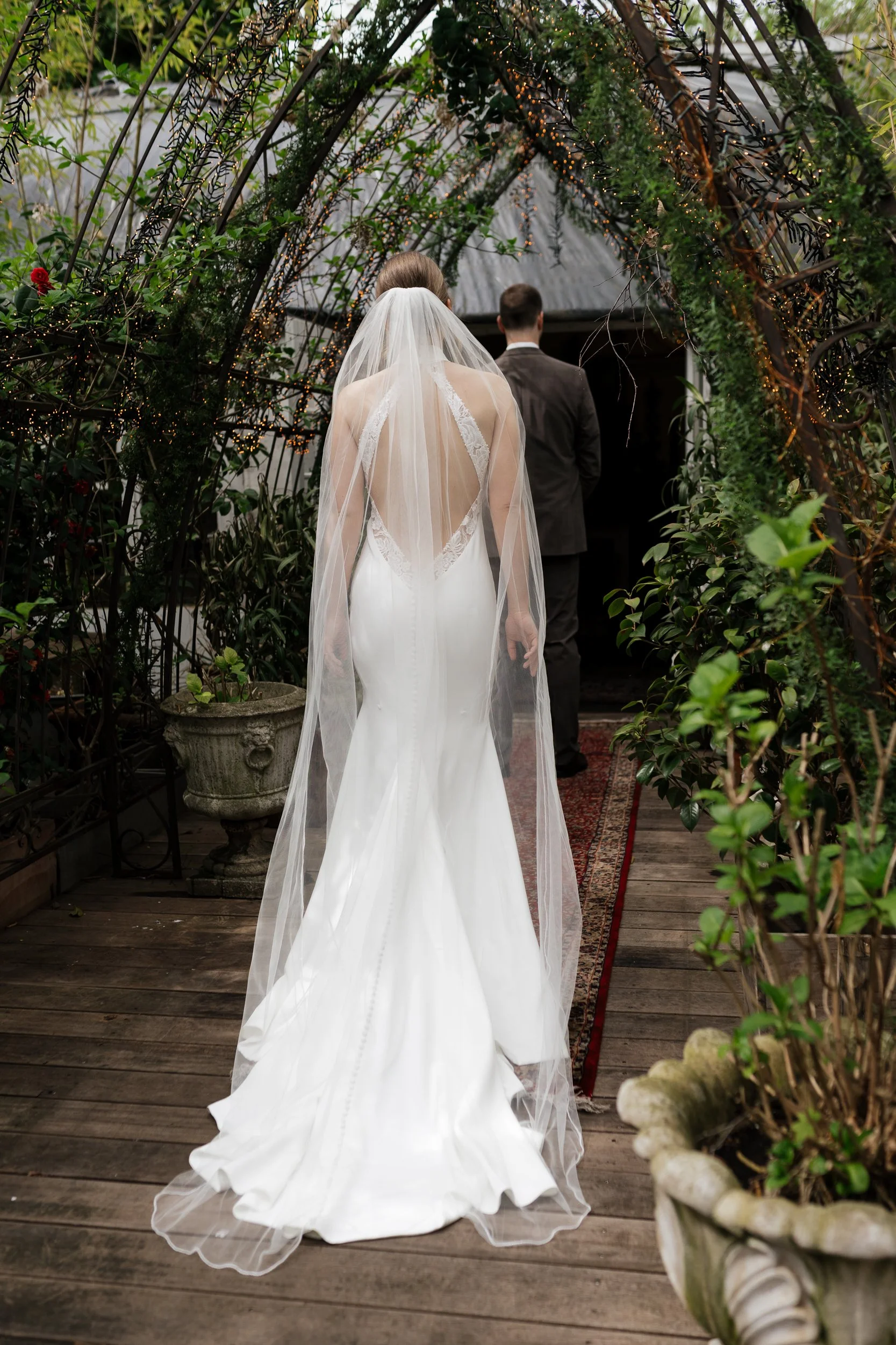 Ein Brautpaar bei einer Hochzeit, die Braut trägt ein weißes Hochzeitskleid und einen Schleier, der Bräutigam trägt einen dunklen Anzug. Sie gehen durch einen Torbogen aus Holz und Lichterketten, umgeben von grünen Pflanzen.