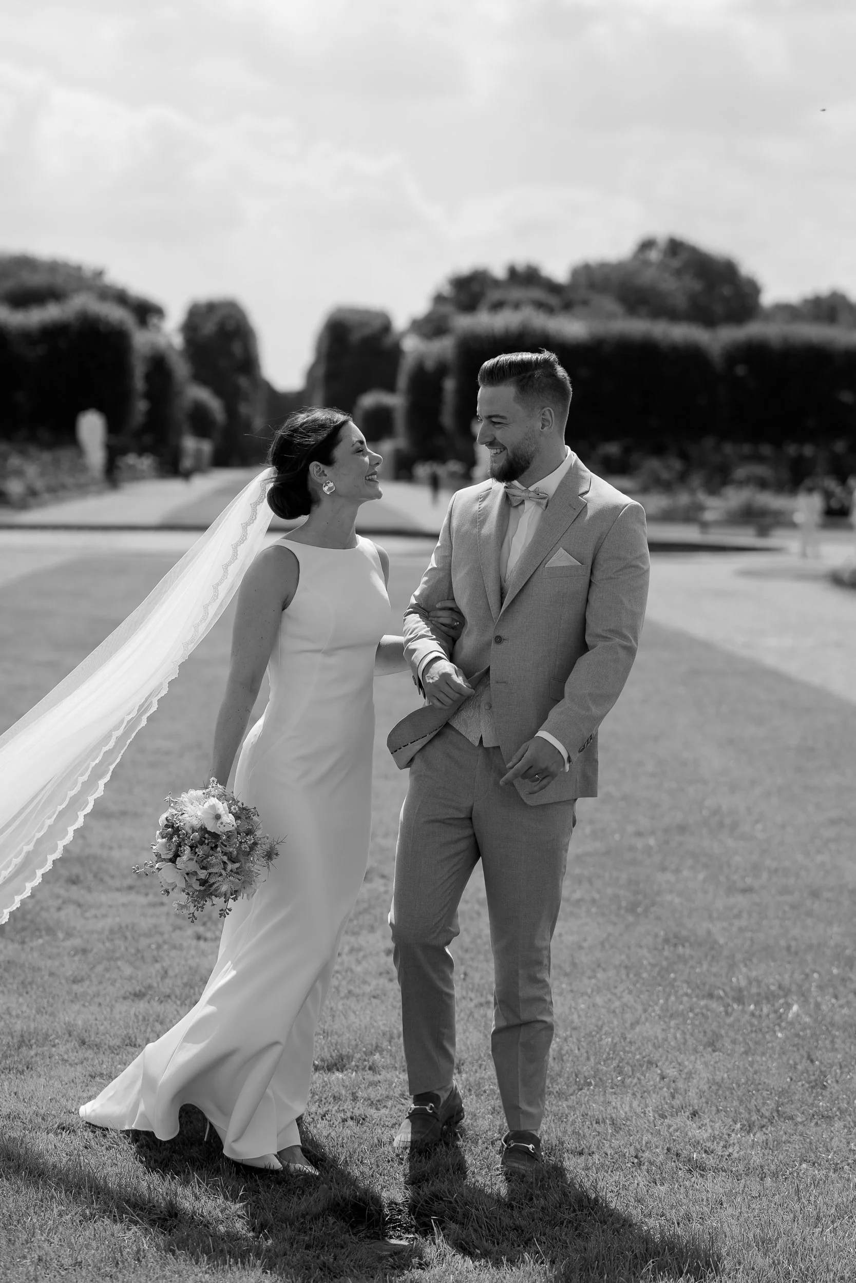 Braut und Bräutigam lachen sich lovend an, auf einer Wiese im Freien bei einer Hochzeit, schwarz-weiß Fotografie.