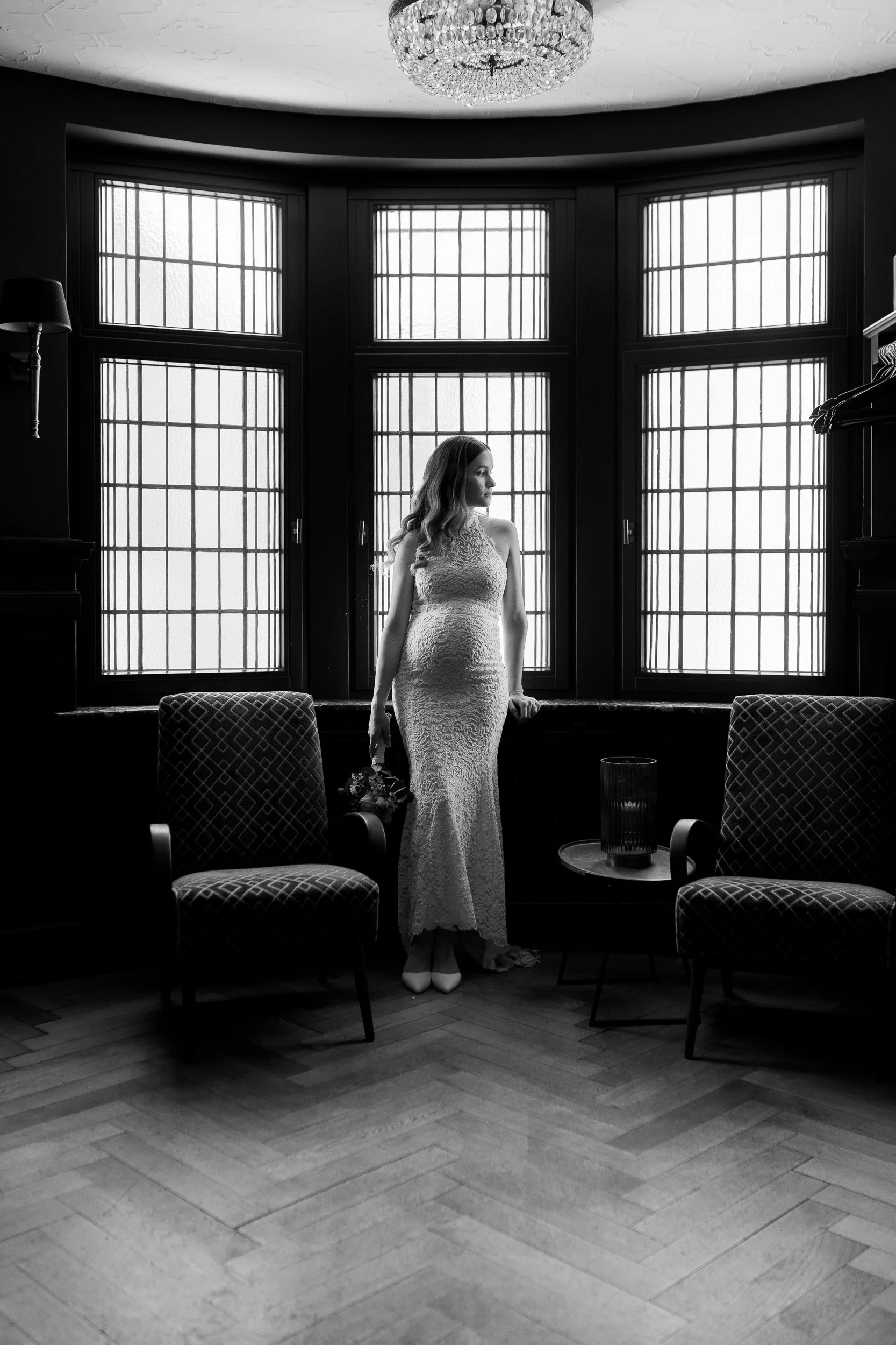 Eine schwarz-weiße Aufnahme einer Frau in einem eleganten Kleid, die in einem Raum vor einem großen Fenster steht. Der Raum ist mit antiken Möbeln und einem Kronleuchter geschmückt.