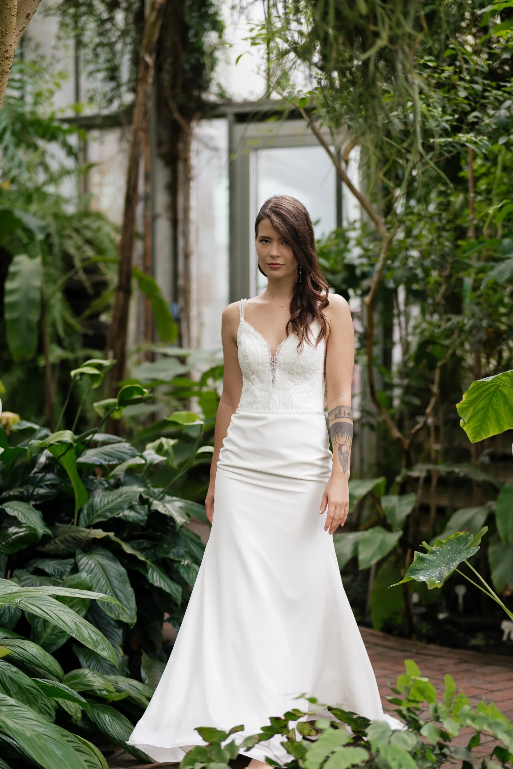 Eine Frau in einem weißen Brautkleid steht in einem grünen Gewächshaus.