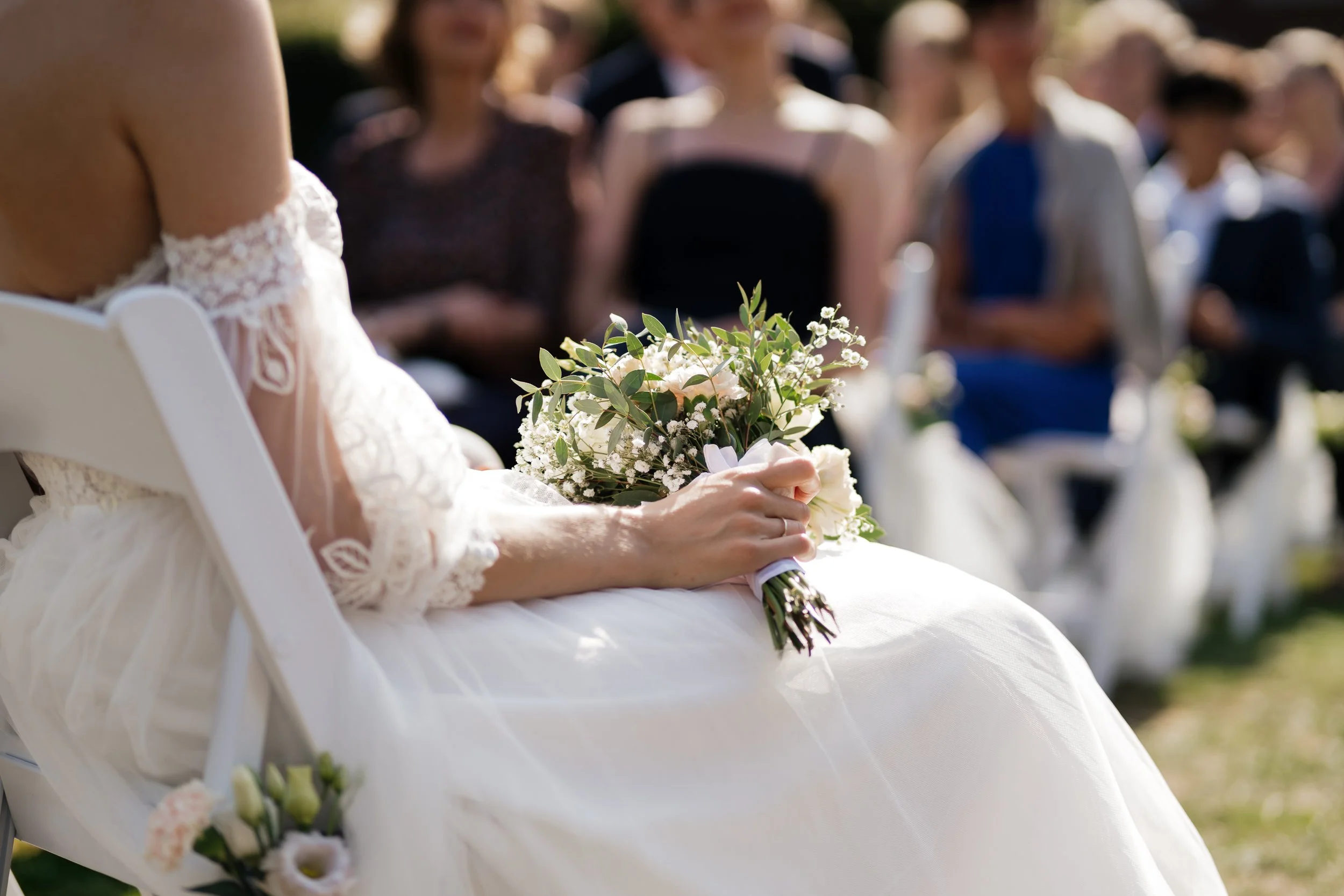 Eine Frau in einem weißen Hochzeitskleid hält einen Blumenstrauß bei einer Hochzeitsfeier im Freien, im Hintergrund sind Gäste sichtbar.