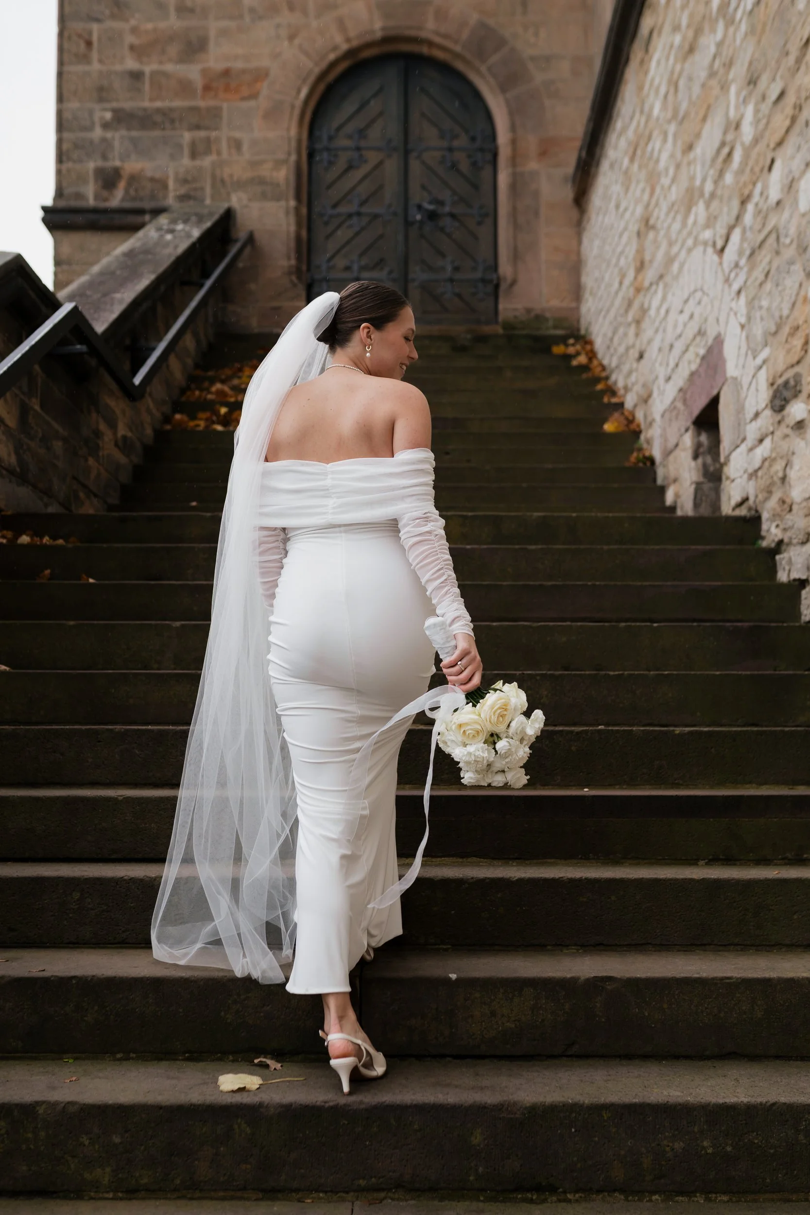 Eine lächelnde Braut in einem weißen Brautkleid mit Schleier, die auf einer Steintreppe vor einem alten Steingebäude steht und einen weißen Brautstrauß hält.
