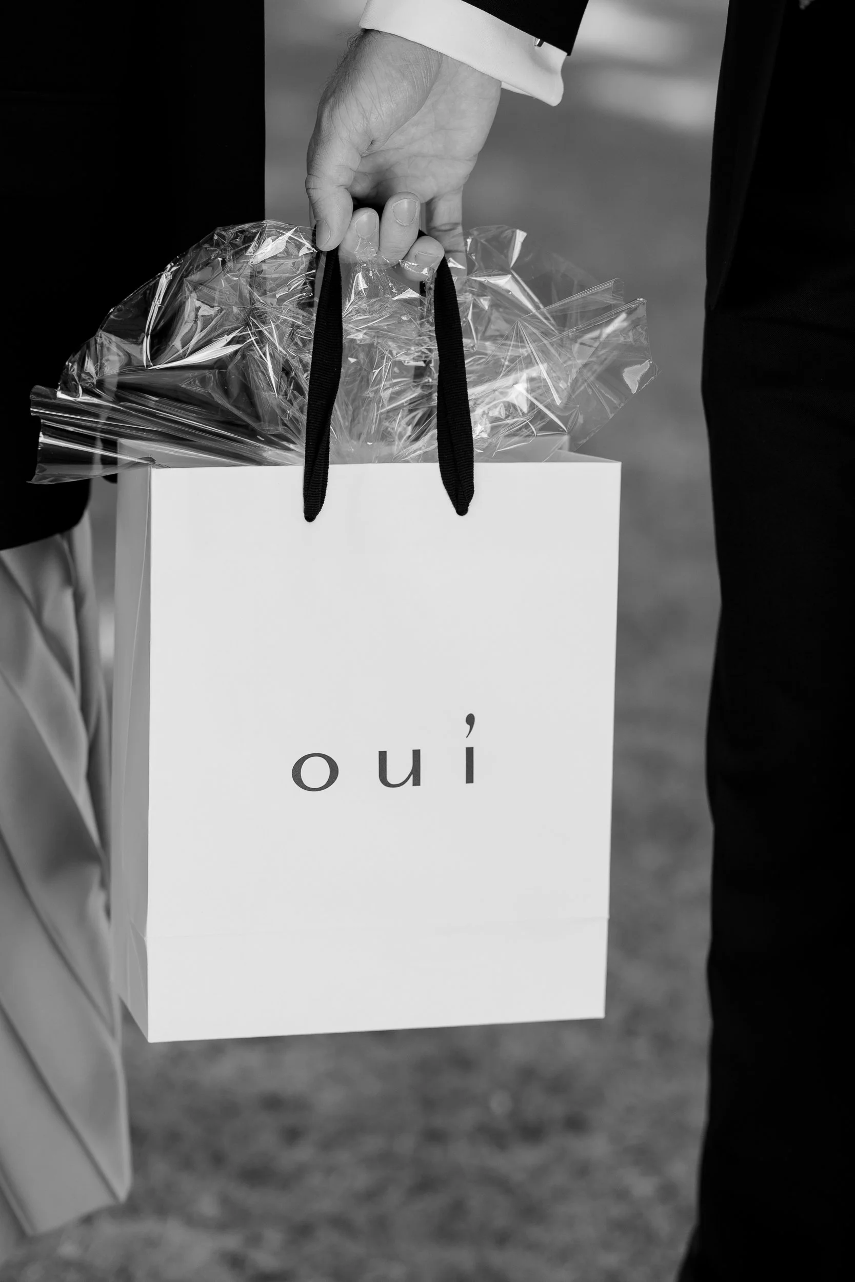 Ein Mensch hält eine weiße Einkaufstüte mit dem Wort 'oui' auf der Vorderseite, wobei eine transparente Geschenkverpackung sichtbar ist.