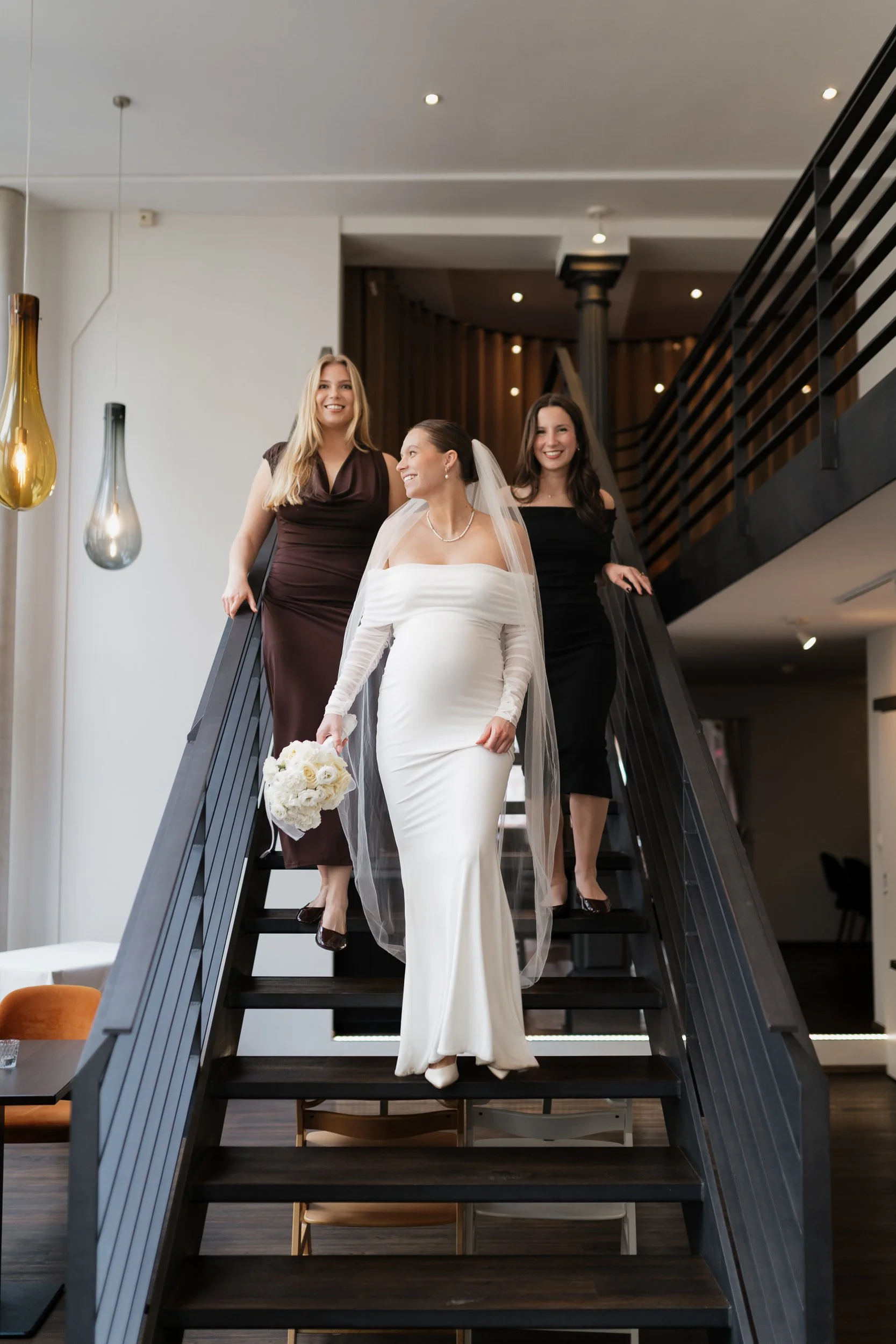 Eine Braut in einem weißen Kleid mit Schleier und Blumenstrauß steigt eine Treppe hinunter, während zwei Frauen in dunklen Kleidern sie begleitet.