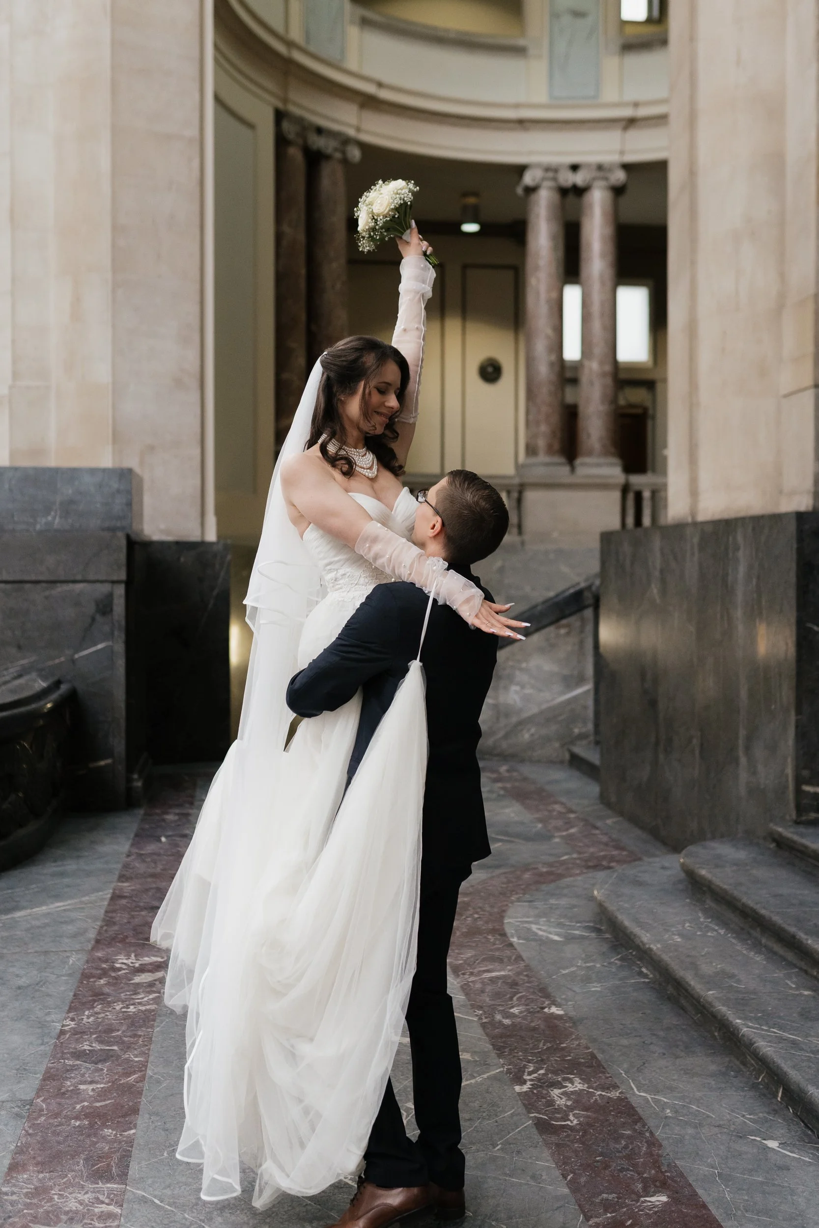 Braut in Hochzeitskleid und Bräutigam in Anzug feiern in einer Kirche, sie hält einen Blumenstrauß hoch, er trägt eine Brille und hebt sie an, bei einer Hochzeit im Innenraum mit Marmorsäulen im Hintergrund.