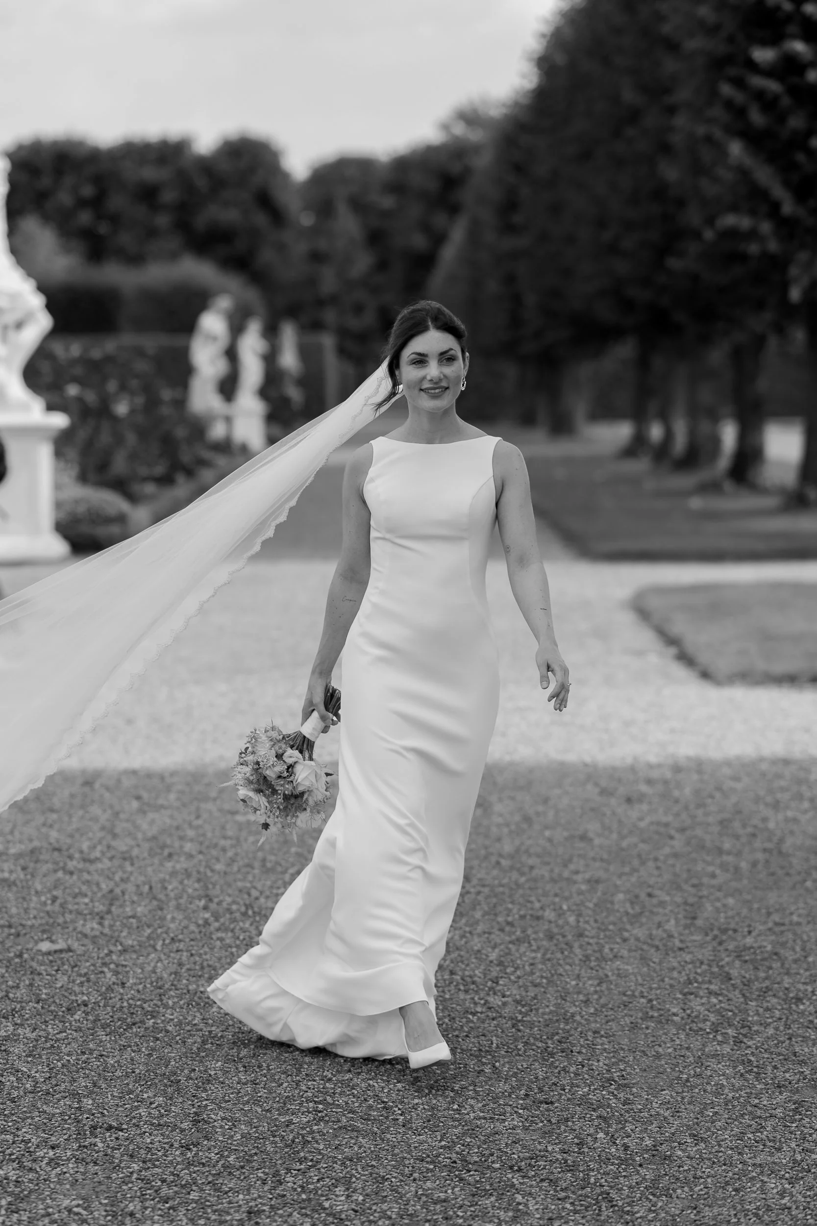 Braut in weißem Kleid mit Blumenstrauß, draußen auf einem Weg, mit Bäumen im Hintergrund, schwarz-weiß Foto.