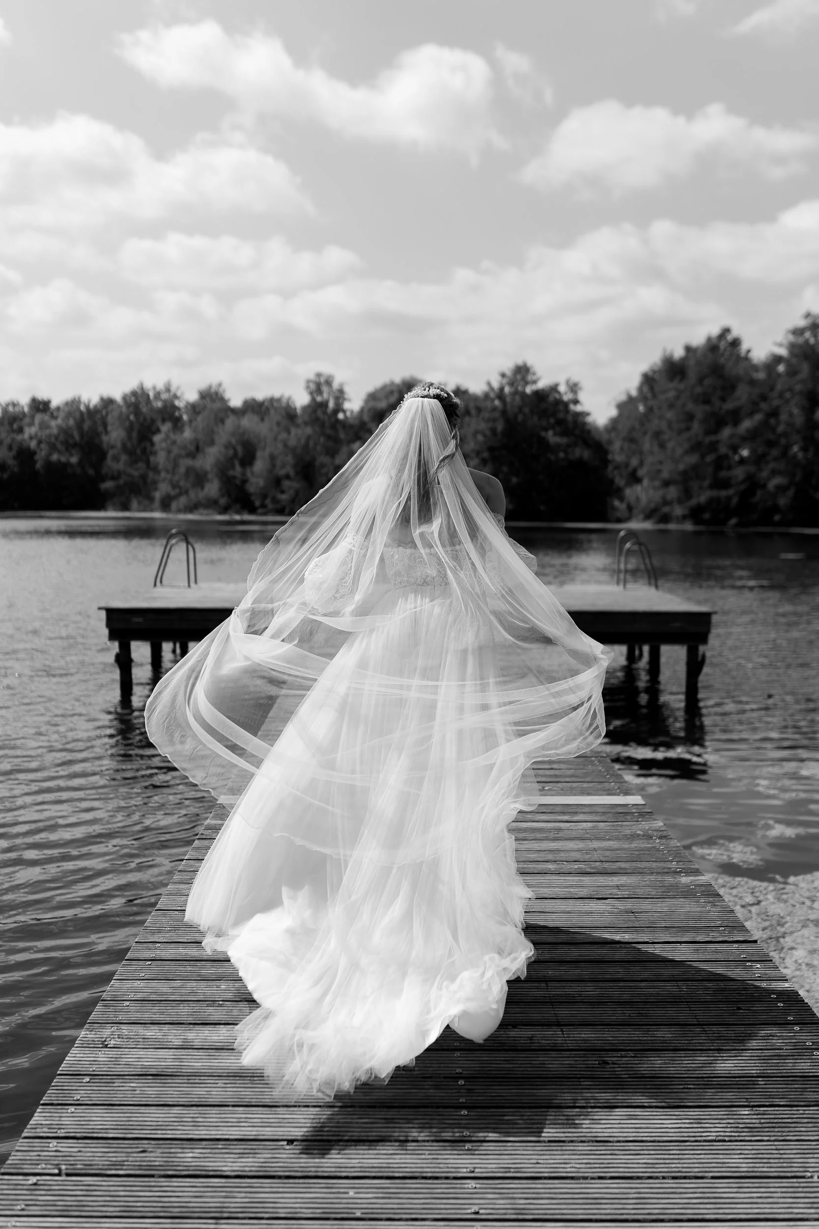 Eine Braut in einem weißen Brautkleid mit Schleier geht auf einem Steg in Richtung Wasser. Im Hintergrund sind Bäume und der Himmel mit Wolken sichtbar, alles in Schwarzweiß.