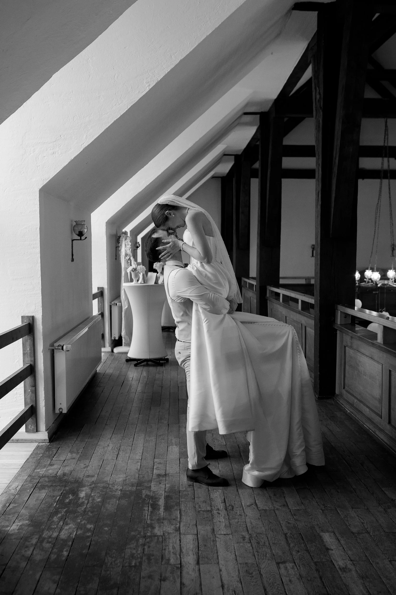 Ein Brautpaar bei einer Hochzeit in einem Raum mit Holzboden, schwarz-weißer Farbgebung, einer schrägen Decke und dekorativen Elementen im Hintergrund. Das Paar befindet sich in einer innigen Umarmung, die Braut trägt ein Hochzeitskleid und die Szene