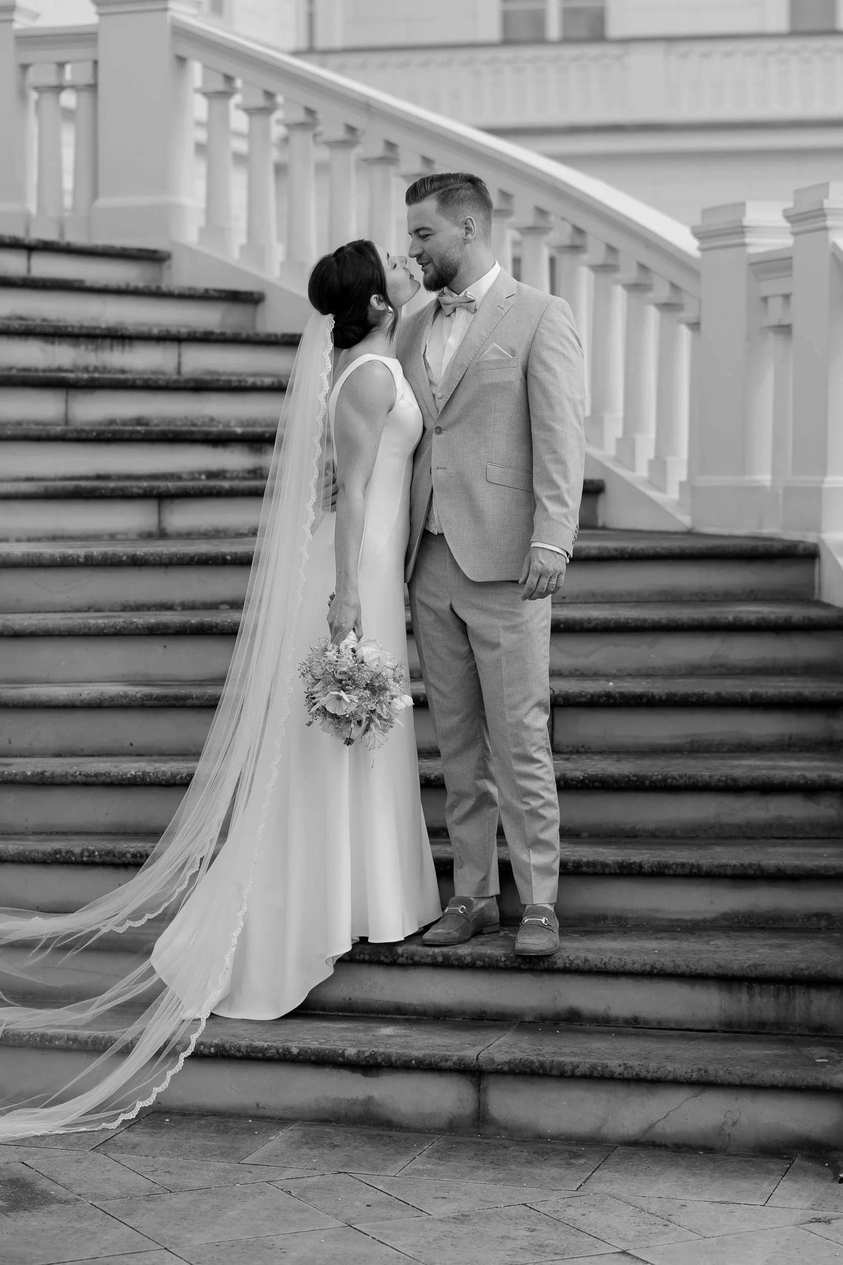 Ein Brautpaar steht auf Treppenstufen, blickt sich liebevoll in die Augen, Tanzhochzeit, schwarz-weiß Aufnahme, elegant gekleidet, romantisches Ambiente.