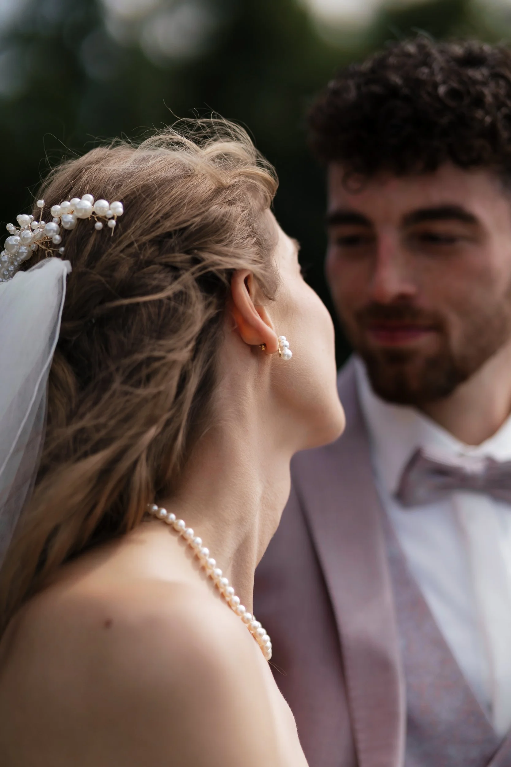 Ein Brautpaar steht sich gegenüber, das Braut trägt ein Perlenhaarkranz und Perlenohrringe, das Bild ist unscharf im Hintergrund. Die Szene wirkt romantisch.