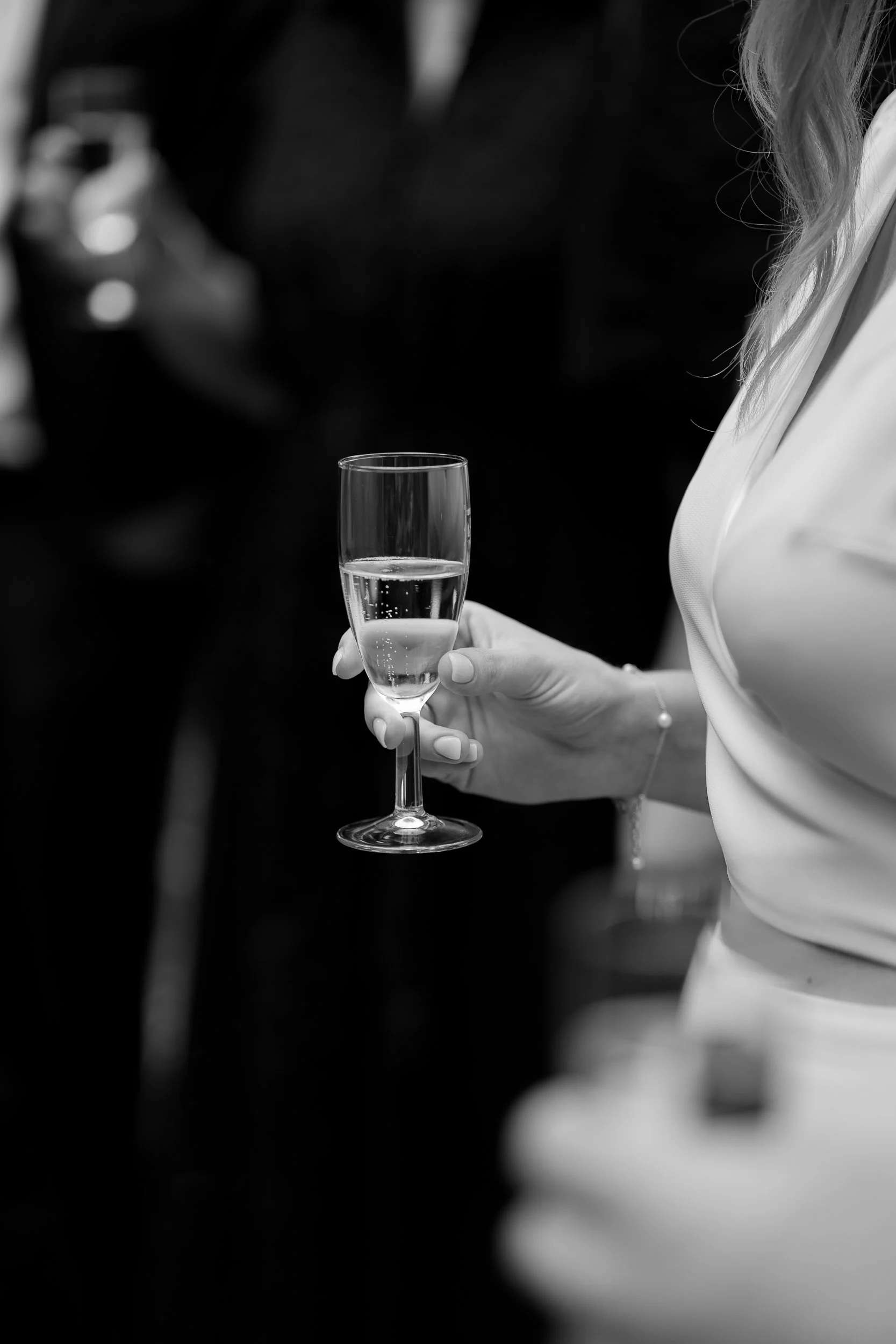 Eine Person hält ein Champagnerglas mit Sekt in der Hand, im Hintergrund sind andere Personen auf einer Party oder Veranstaltung zu erkennen.