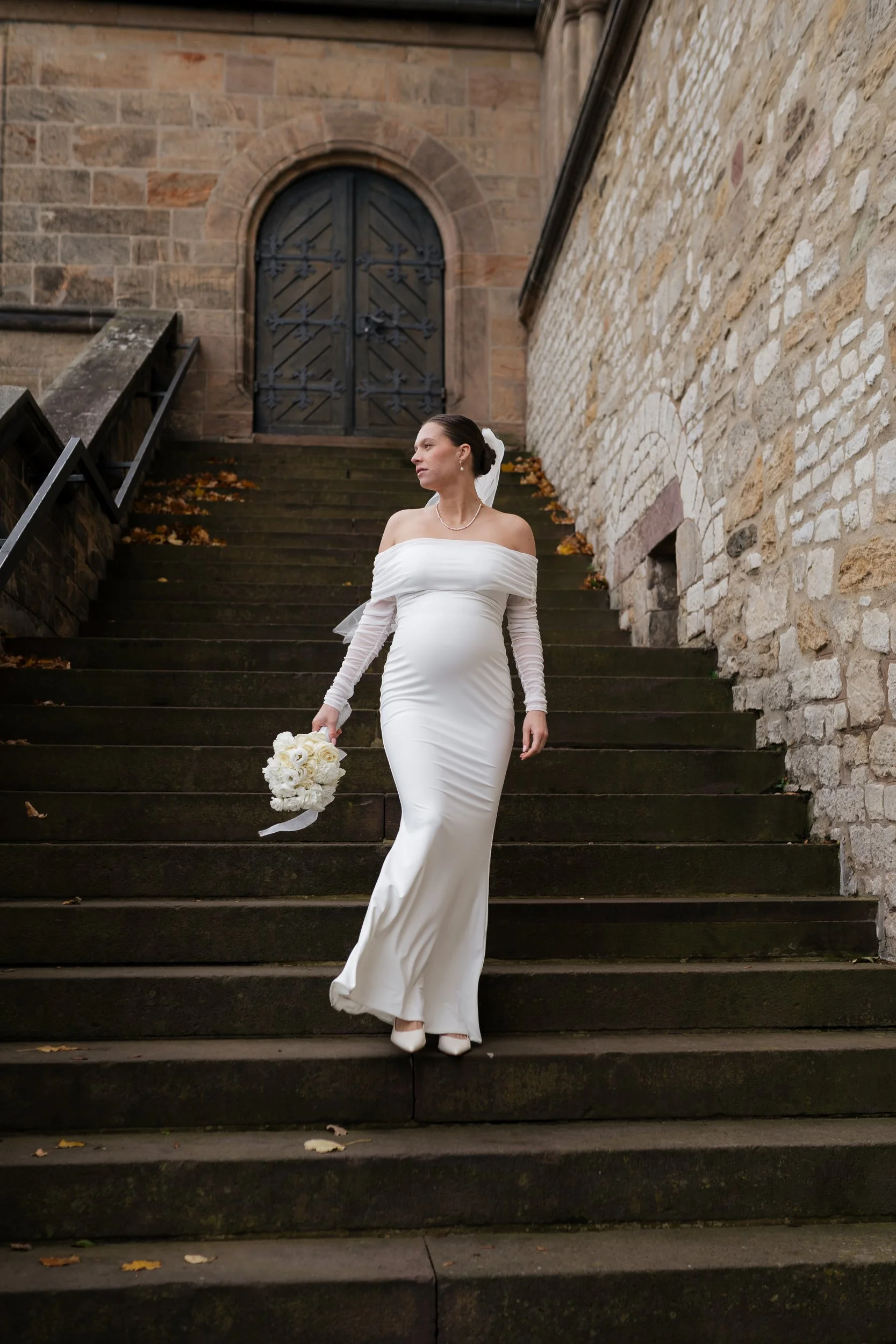 Eine schwangere Frau in einem weißen Brautkleid, die auf einer alten Steintreppe vor einer Steinmauer steht, hält einen Blumenstrauß in der Hand und schaut nach rechts.
