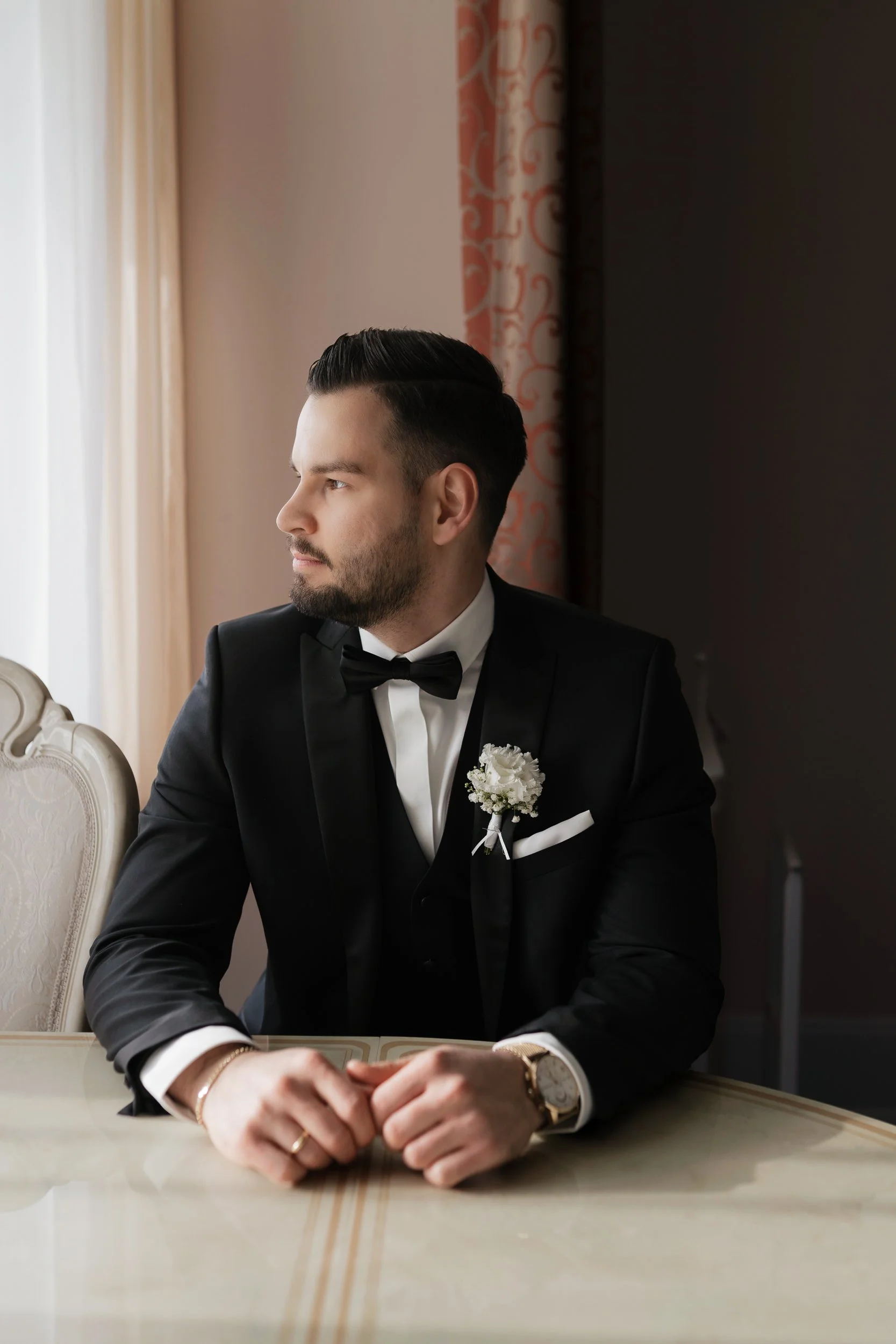 Ein gut gekleideter Mann in einem schwarzen Anzug, weißem Hemd und Fliege, sitzt an einem Tisch in einem eleganten Raum mit weißen und pastellfarbenen Vorhängen, betrachtet nach links und trägt eine goldene Uhr und Schmuck.