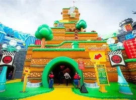 A large, colorful toy-themed structure resembling a mountain with green trees, resembling a scene from a video game or play area, with two children entering and adults nearby.