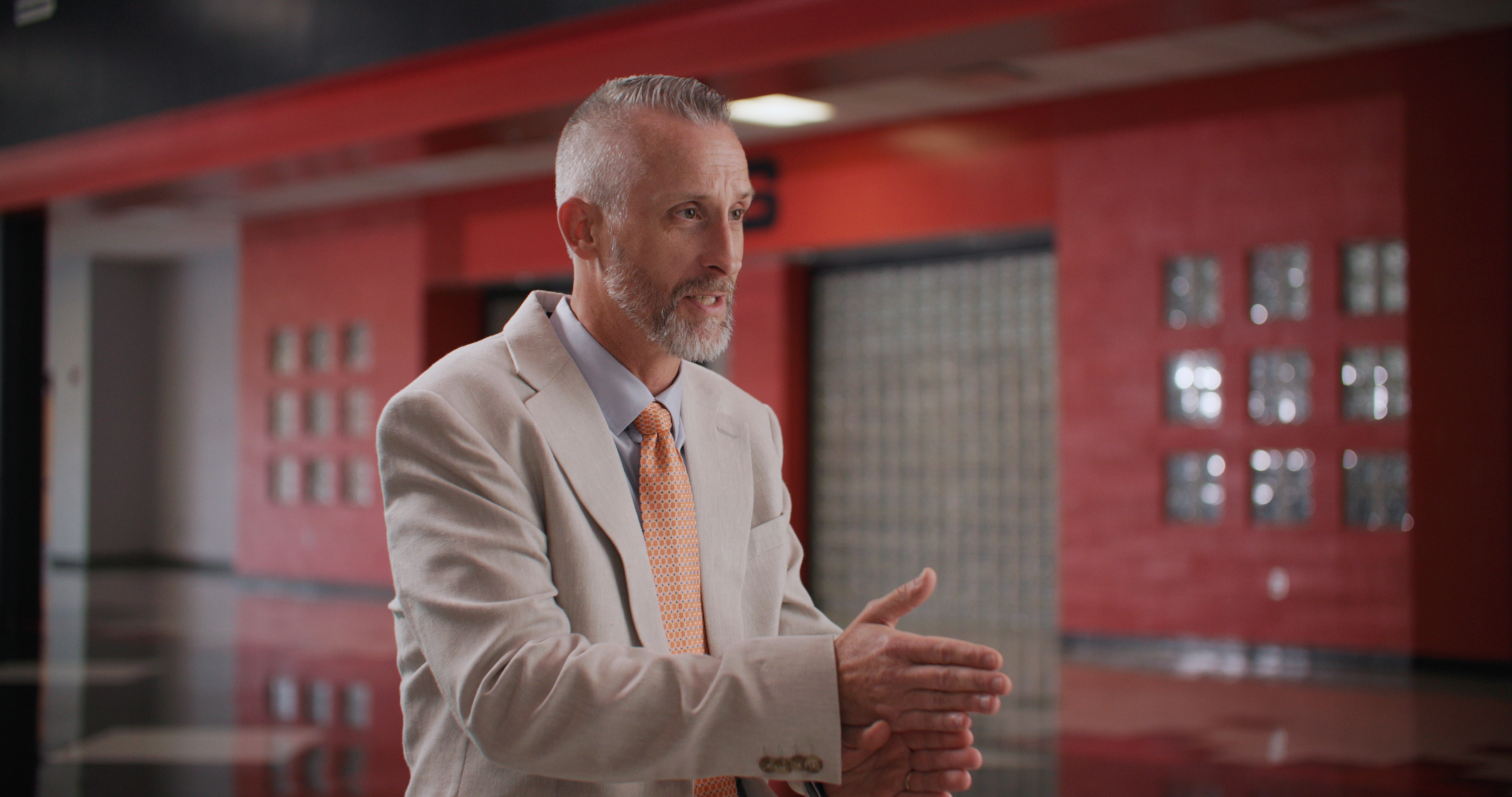A middle-aged man with gray hair and a beard, dressed in a beige suit and orange patterned tie, is talking and gesturing with his hands indoors against a red wall with glass blocks.
