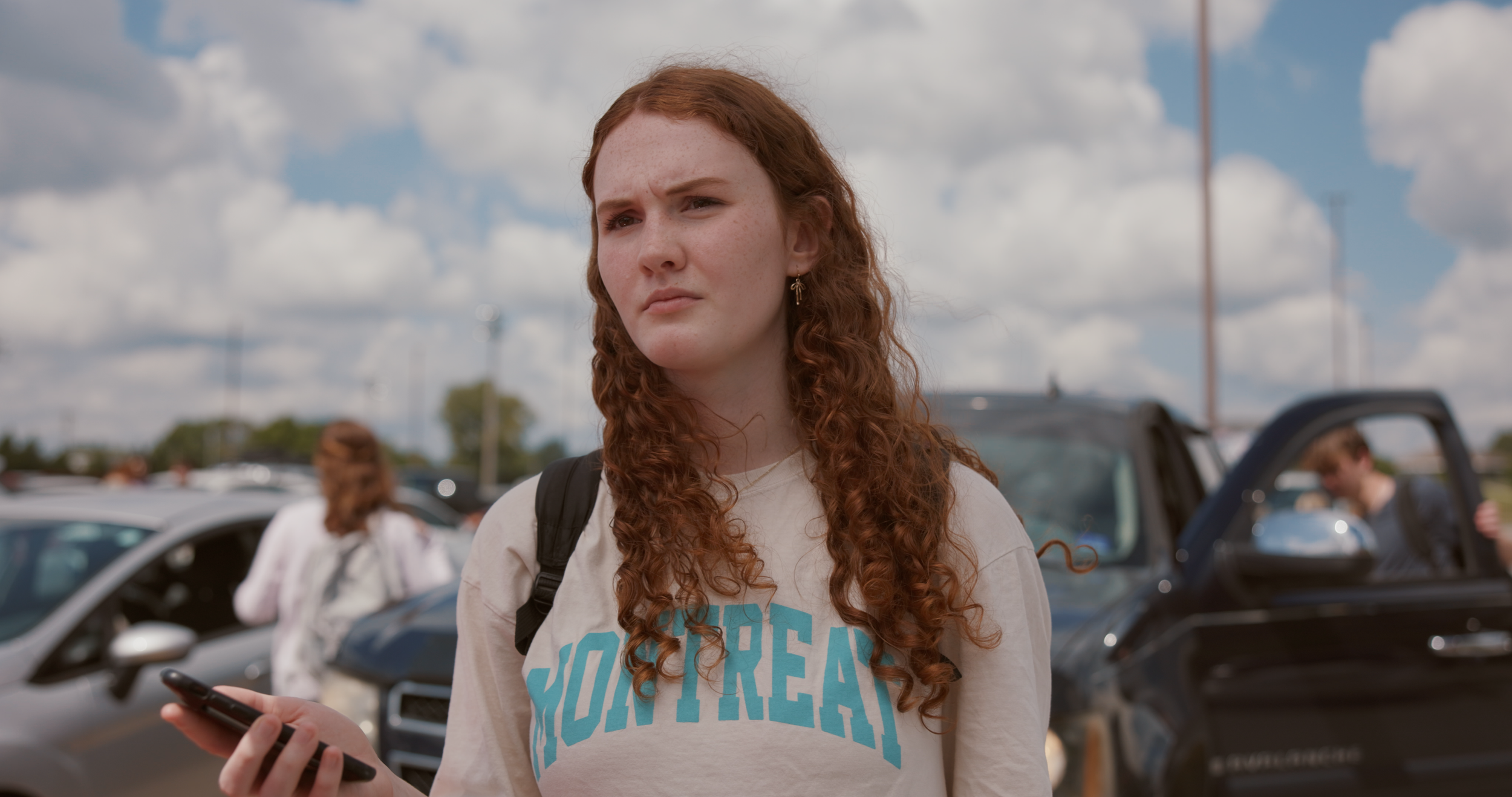 A young woman with curly red hair holding a phone, standing in a parking lot on a partly cloudy day. She wears a beige sweatshirt with blue lettering, a backpack, and earrings. There's a black pickup truck and other cars in the background, with a per