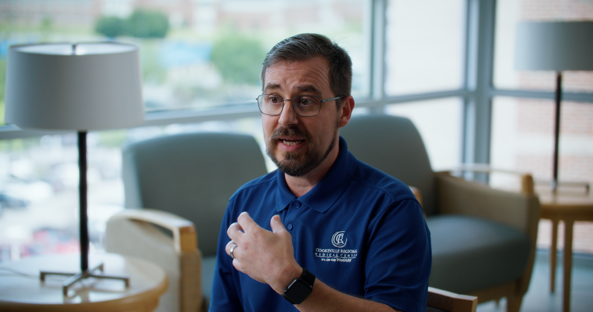 A man wearing glasses and a blue polo shirt with 'Cookeville Regional Medical Center' logo, sitting in a waiting area, gesturing with his right hand on his chest, speaking, with lamps and chairs in the background.