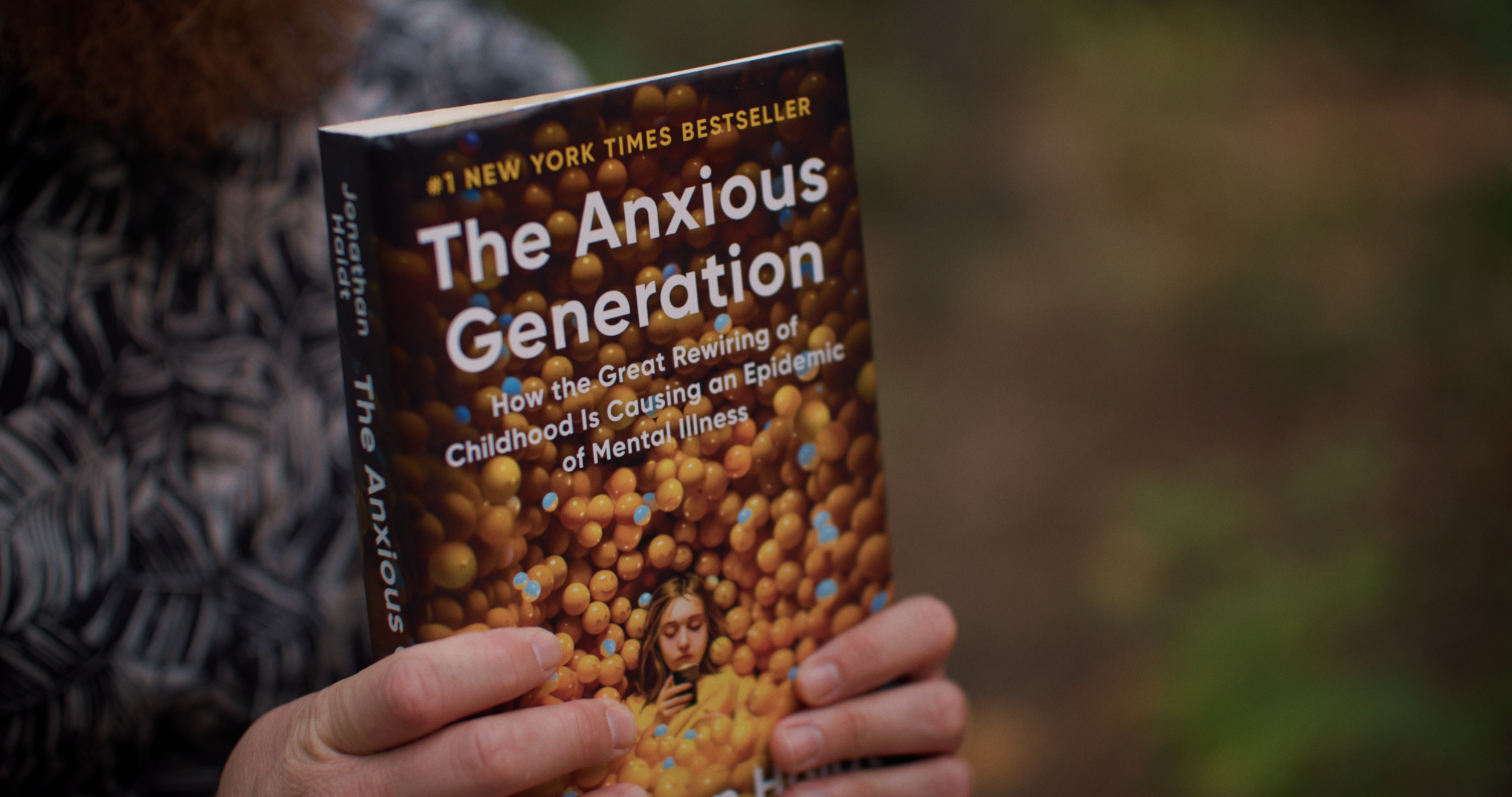 A person holding a book titled "The Anxious Generation" with a background of brown and blue beads and a small photo of a girl looking at her phone.