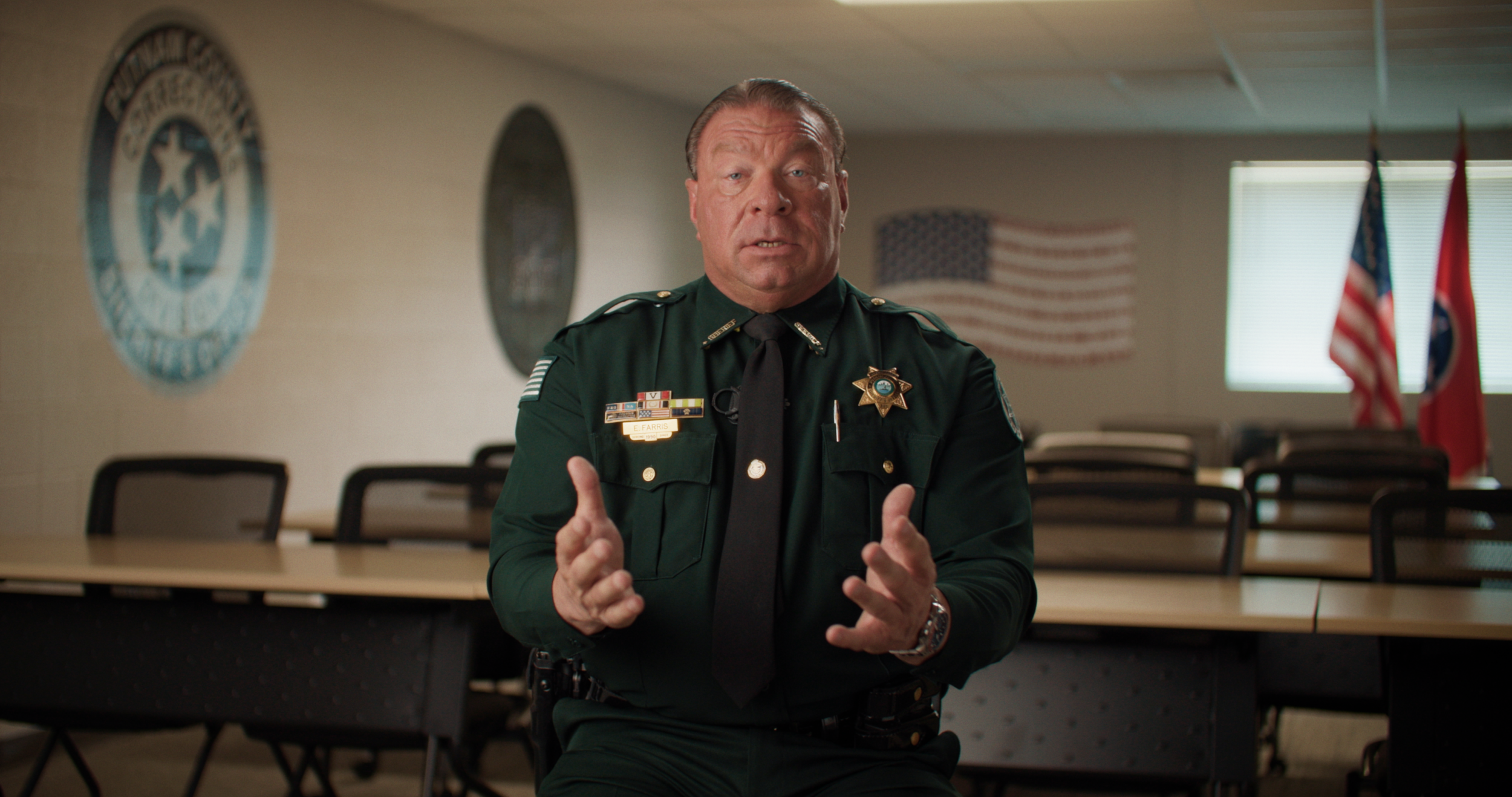 A police officer in uniform is sitting in a meeting room, speaking and gesturing with his hands. The room has American flags and police emblems on the walls.