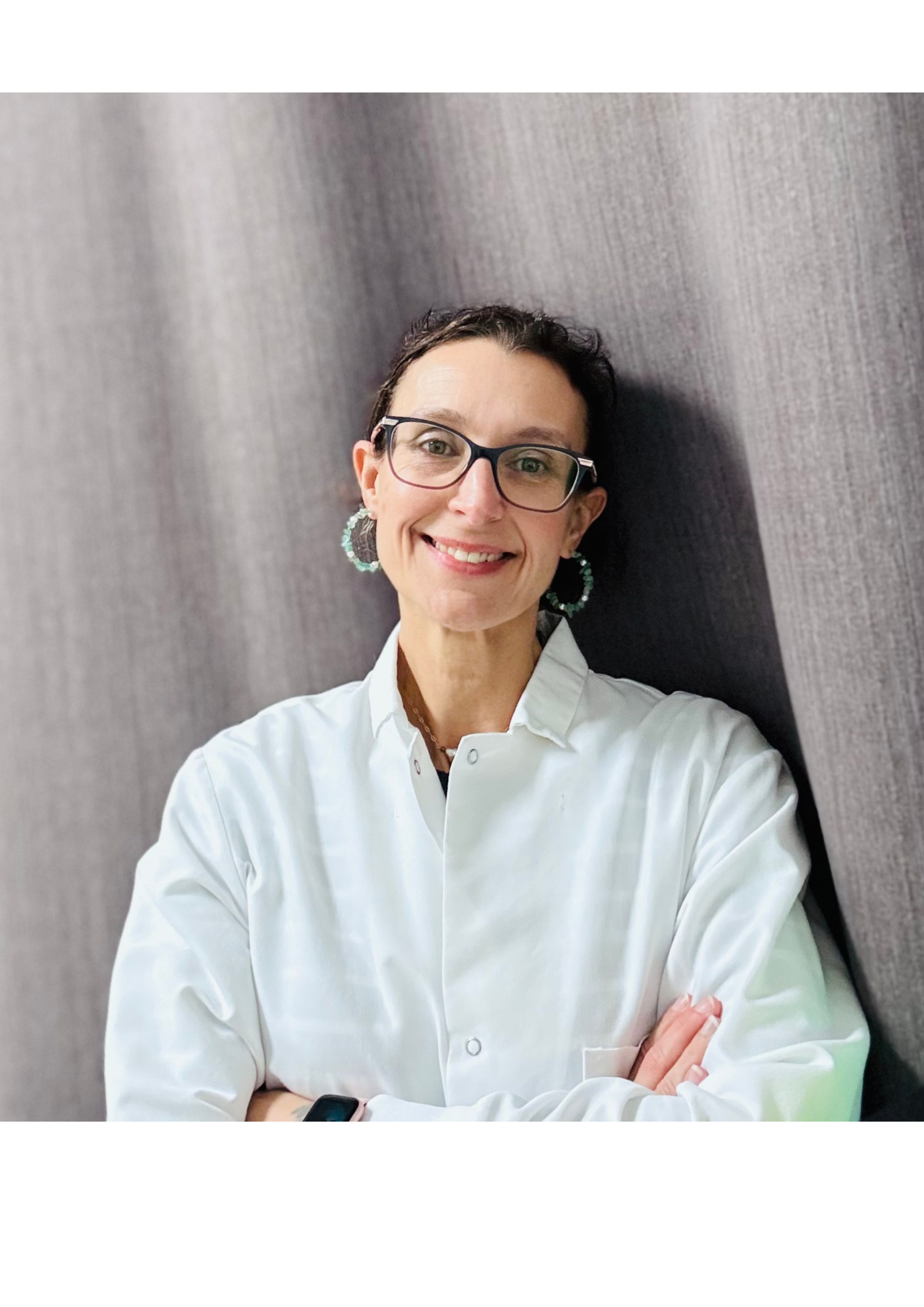 A woman with short dark hair, glasses, and hoop earrings, wearing a white lab coat, standing against a textured gray wall, smiling with arms crossed.
