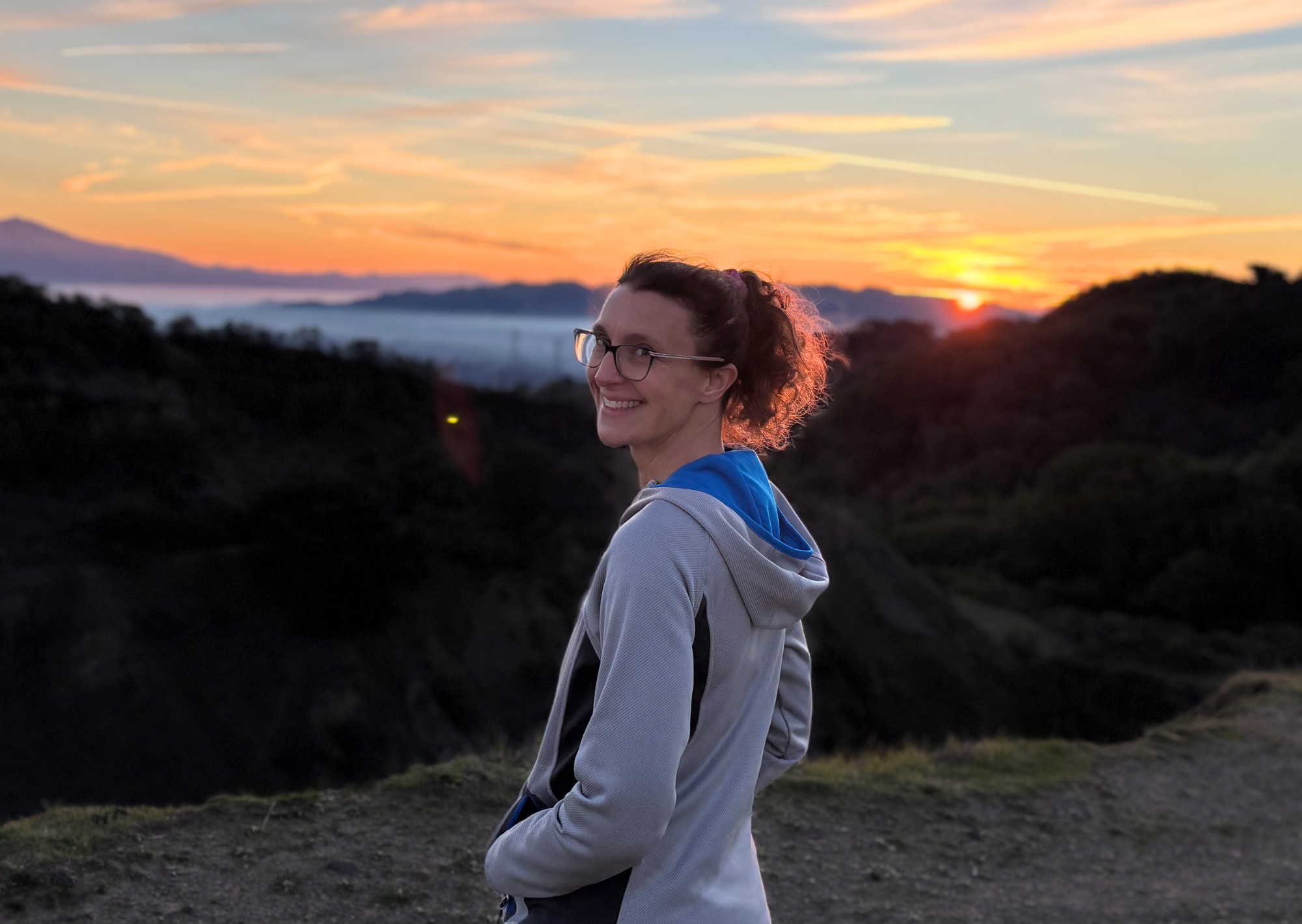 A woman with glasses and curly hair smiling at sunset, standing outdoors on a hill with a landscape of mountains and a colorful sky in the background.