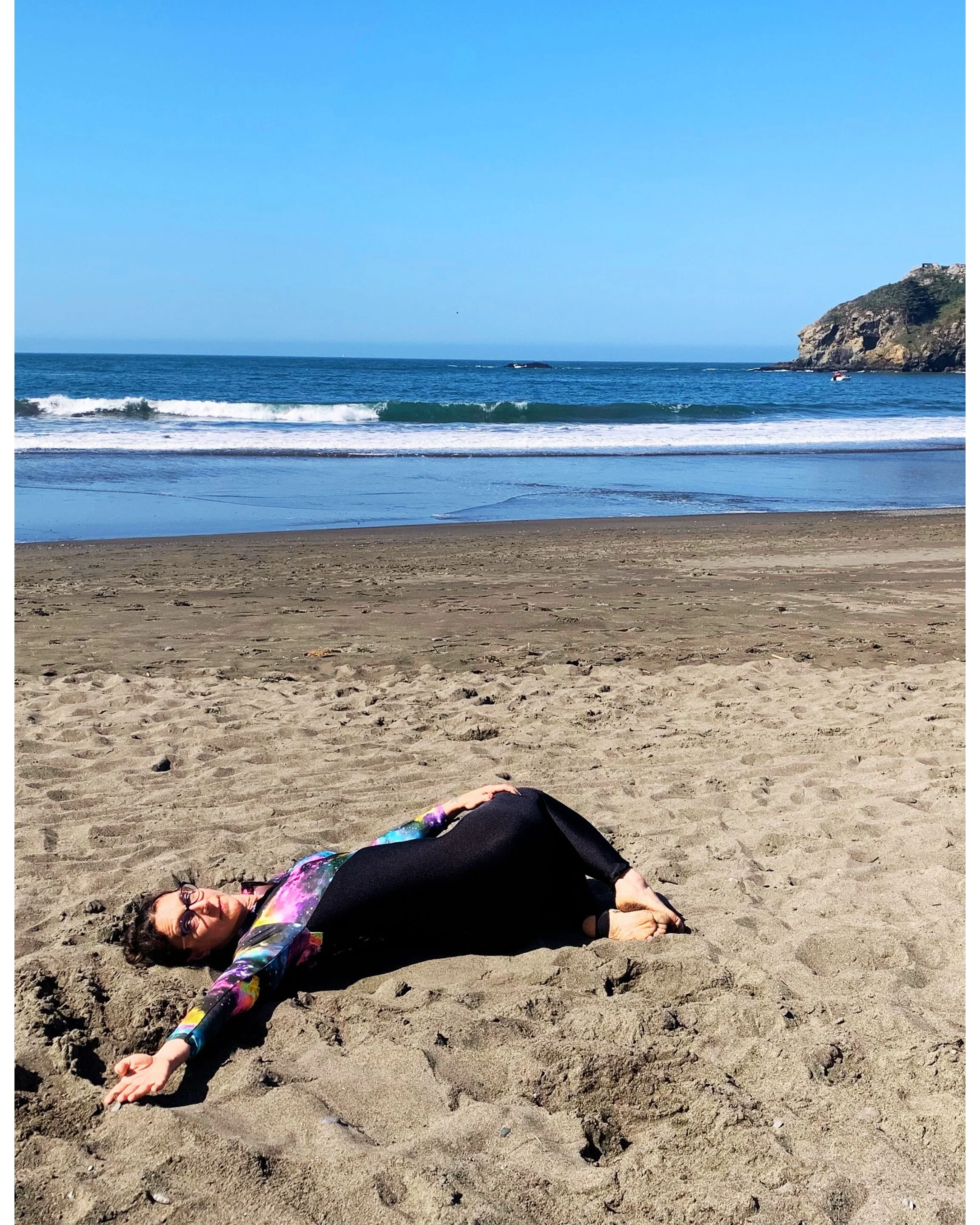 Woman lying on the sandy beach, wearing sunglasses and a colorful long-sleeve top with black pants, with ocean waves and a cliff in the background.
