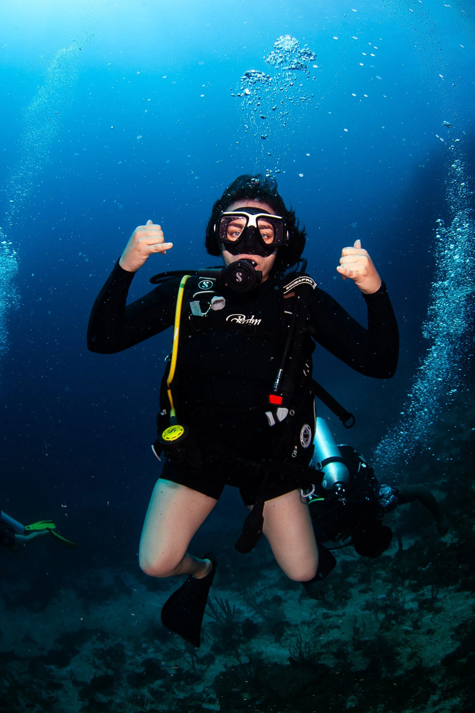 Person scuba diving underwater, wearing a wetsuit, mask, and fins, surrounded by blue water and small bubbles.