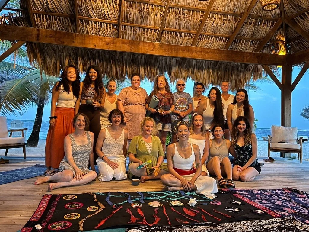 Group of women and men posing under a thatched roof on a beachside deck with the ocean in the background, some sitting on the floor, others standing, with colorful tapestries and crafts in front.