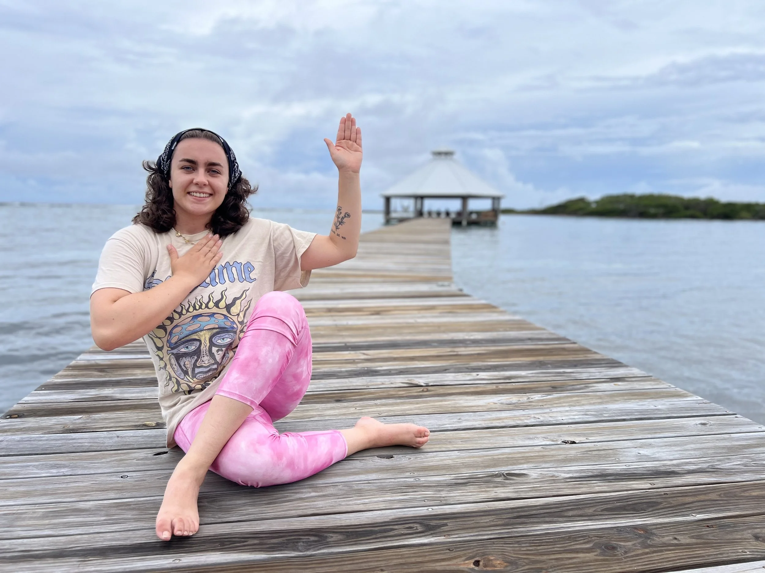 A young woman with shoulder-length dark hair, smiling and wearing a bandana, a graphic T-shirt, and pink tie-dye leggings, sitting on a wooden dock by the water with a small pavilion in the background, gesturing with her right hand raised and her left hand on her chest.