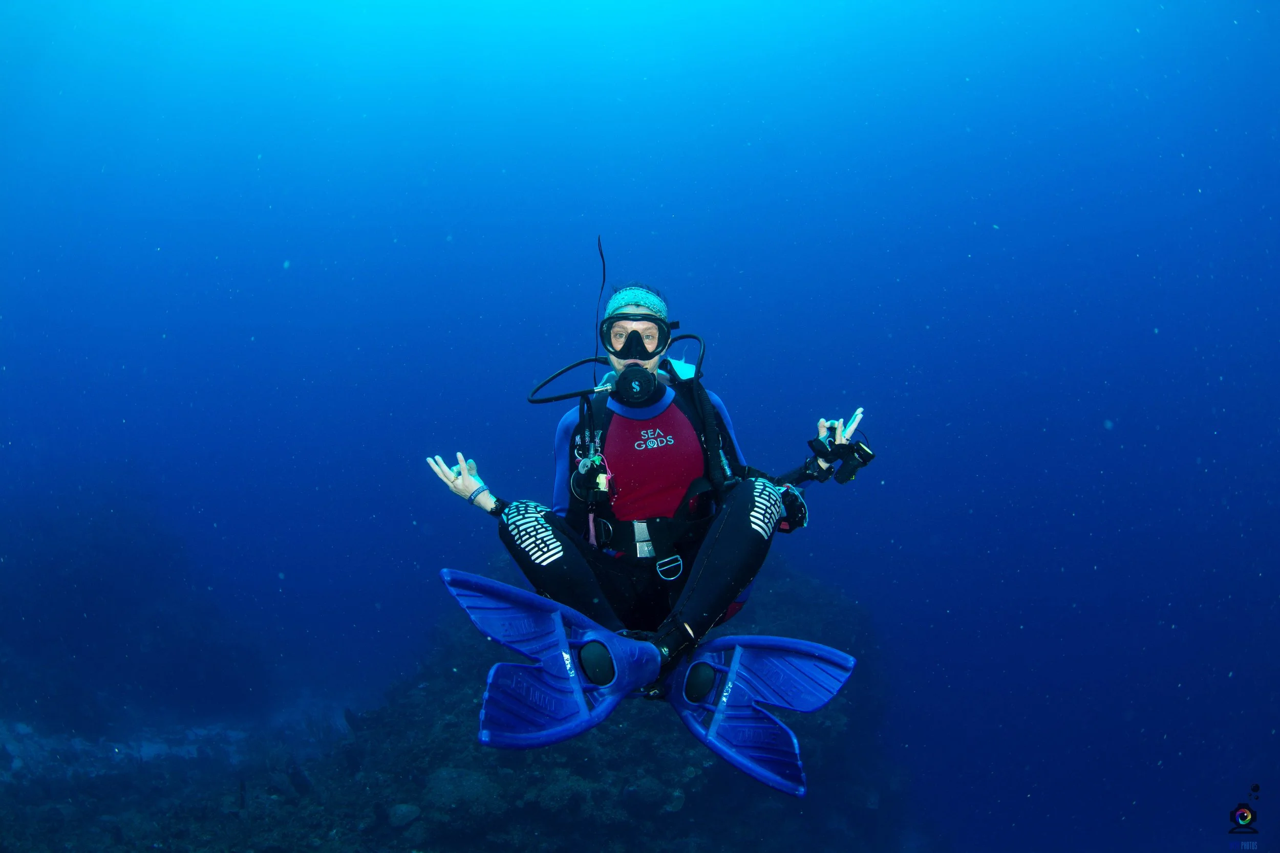 A scuba diver sitting cross-legged underwater, making peaceful hand gestures, equipped with a diving mask, regulator, and bright blue fins, against a deep blue ocean background.