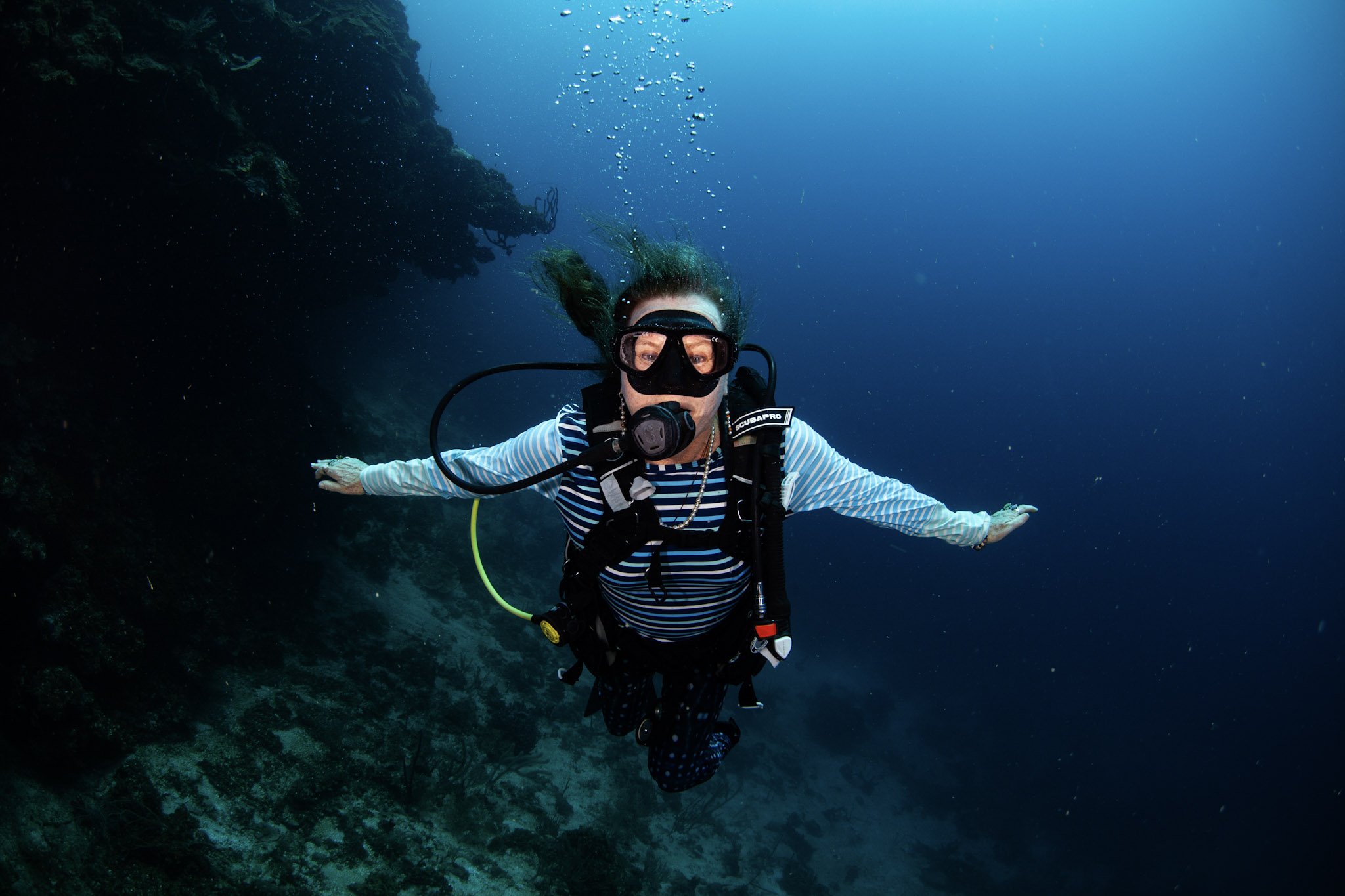 Woman scuba diving underwater with arms extended, wearing a striped shirt, mask, and scuba gear.