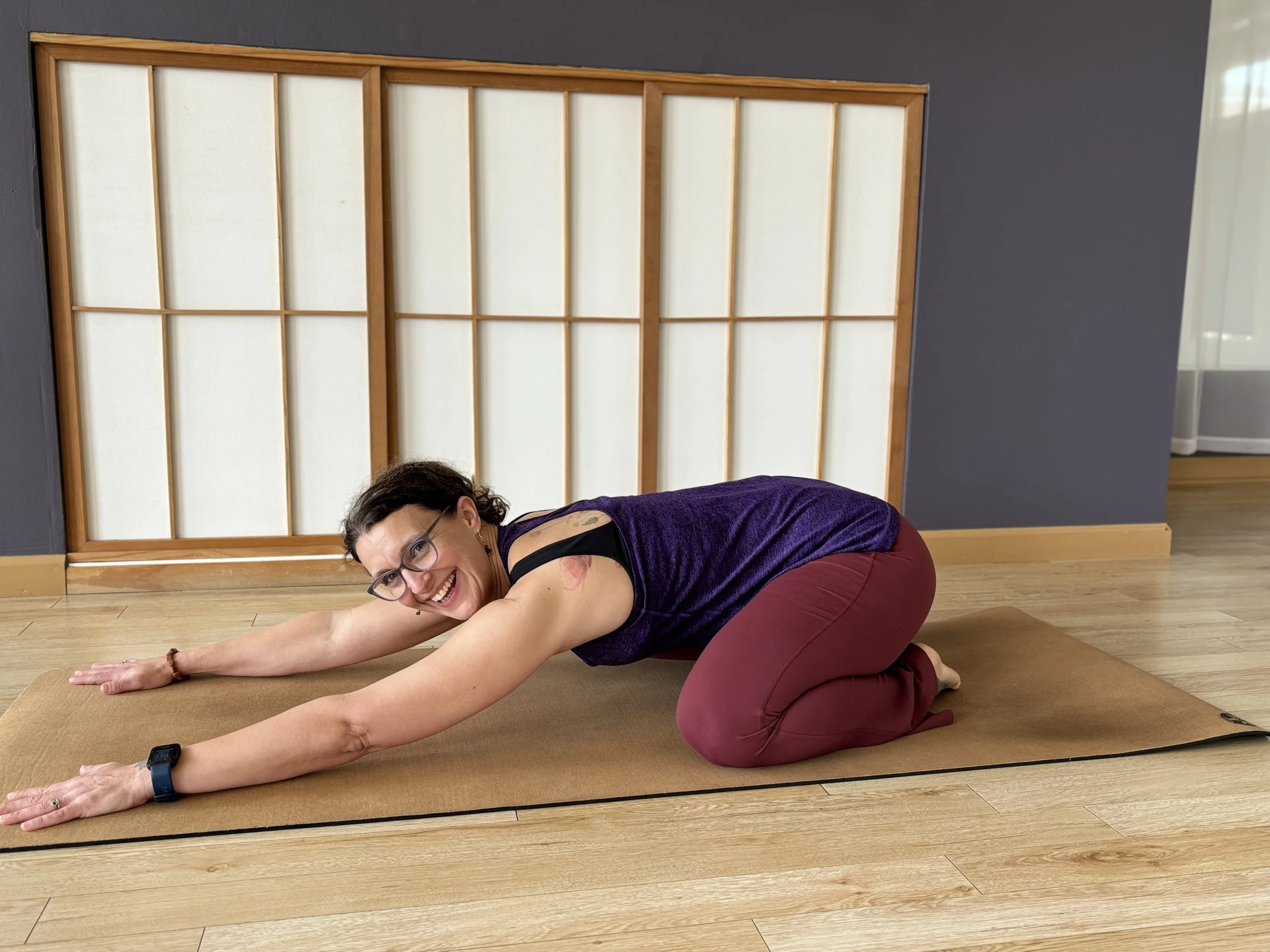 A woman with glasses and short dark hair practicing yoga on a mat, in a kneeling forward bend position, smiling while looking at the camera.