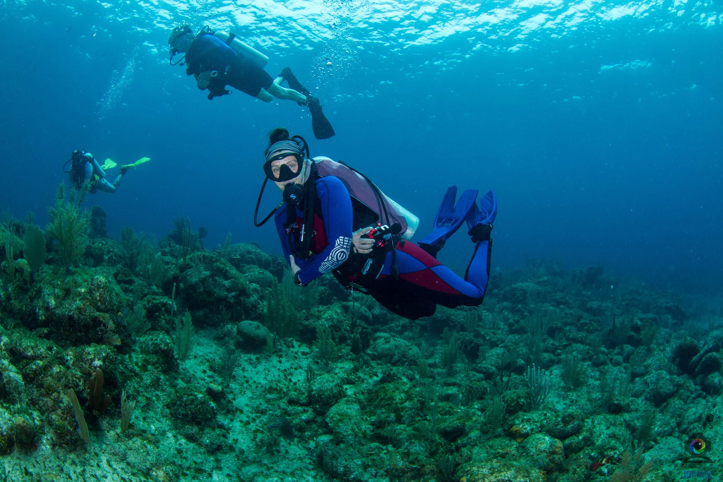 Three scuba divers exploring coral reef underwater. One woman in the foreground wearing blue wetsuit, mask, and fins, holding a camera. Two men are farther back, also in scuba gear. Coral and marine life are visible on the ocean floor.