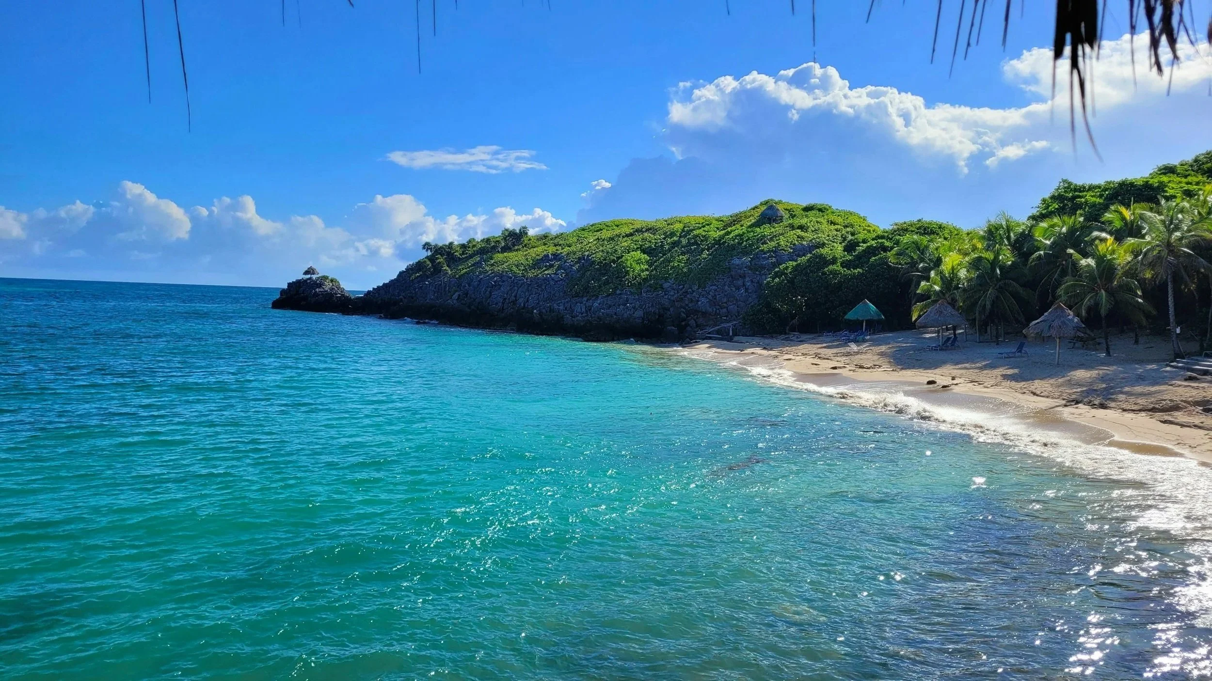 Tropical beach with turquoise water, sandy shore, thatched umbrellas, palm trees, and green hillside under a blue sky with clouds.