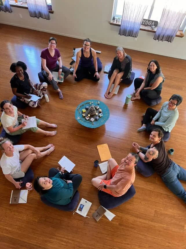 A group of eleven women sitting in a circle on a wooden floor, centered around a blue tray with candles and pinecones, in a room with light-colored walls and windows with blue curtains.