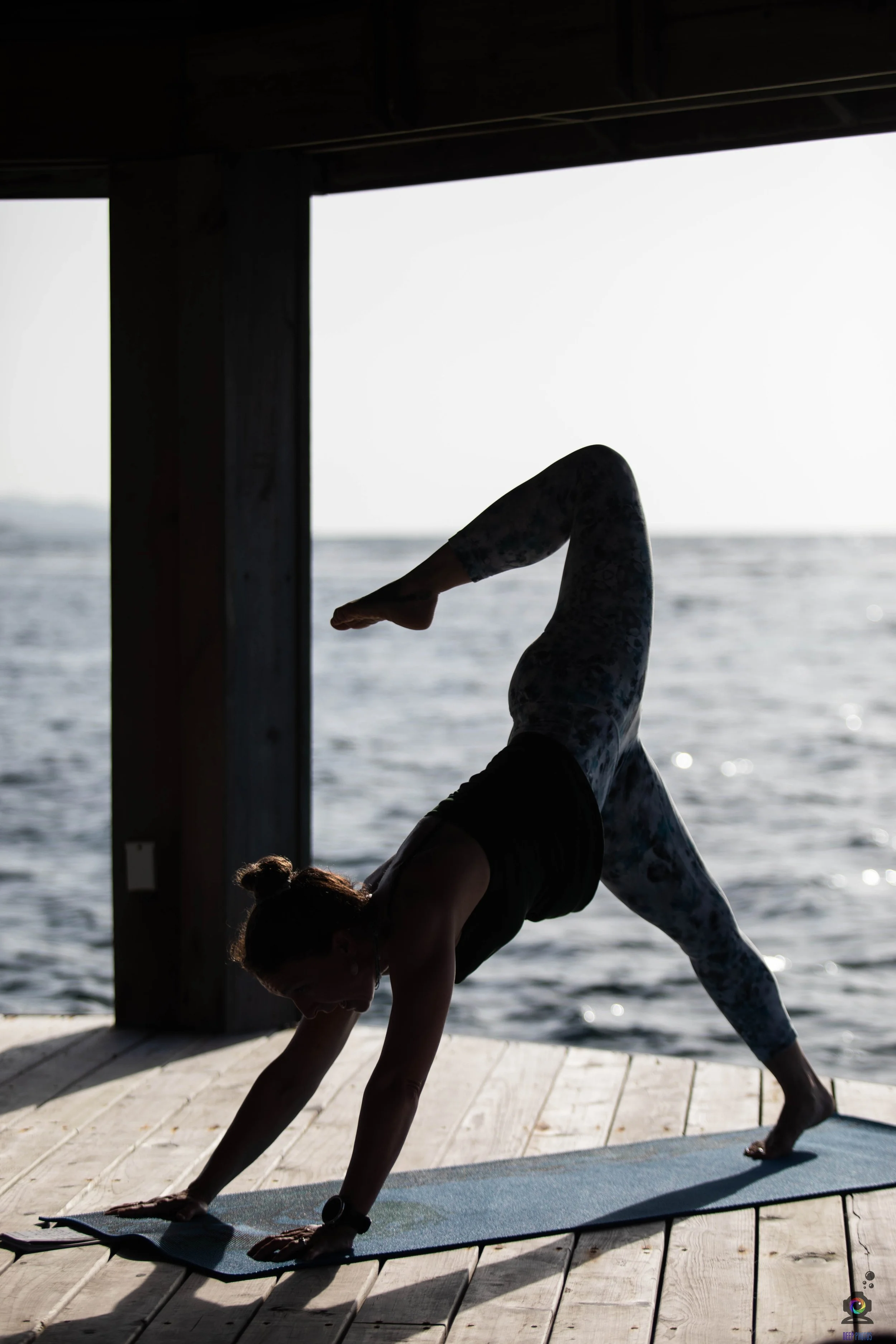 A woman practicing yoga outdoors on a dock by the water, in a downward dog pose with one leg raised, during sunset or sunrise.