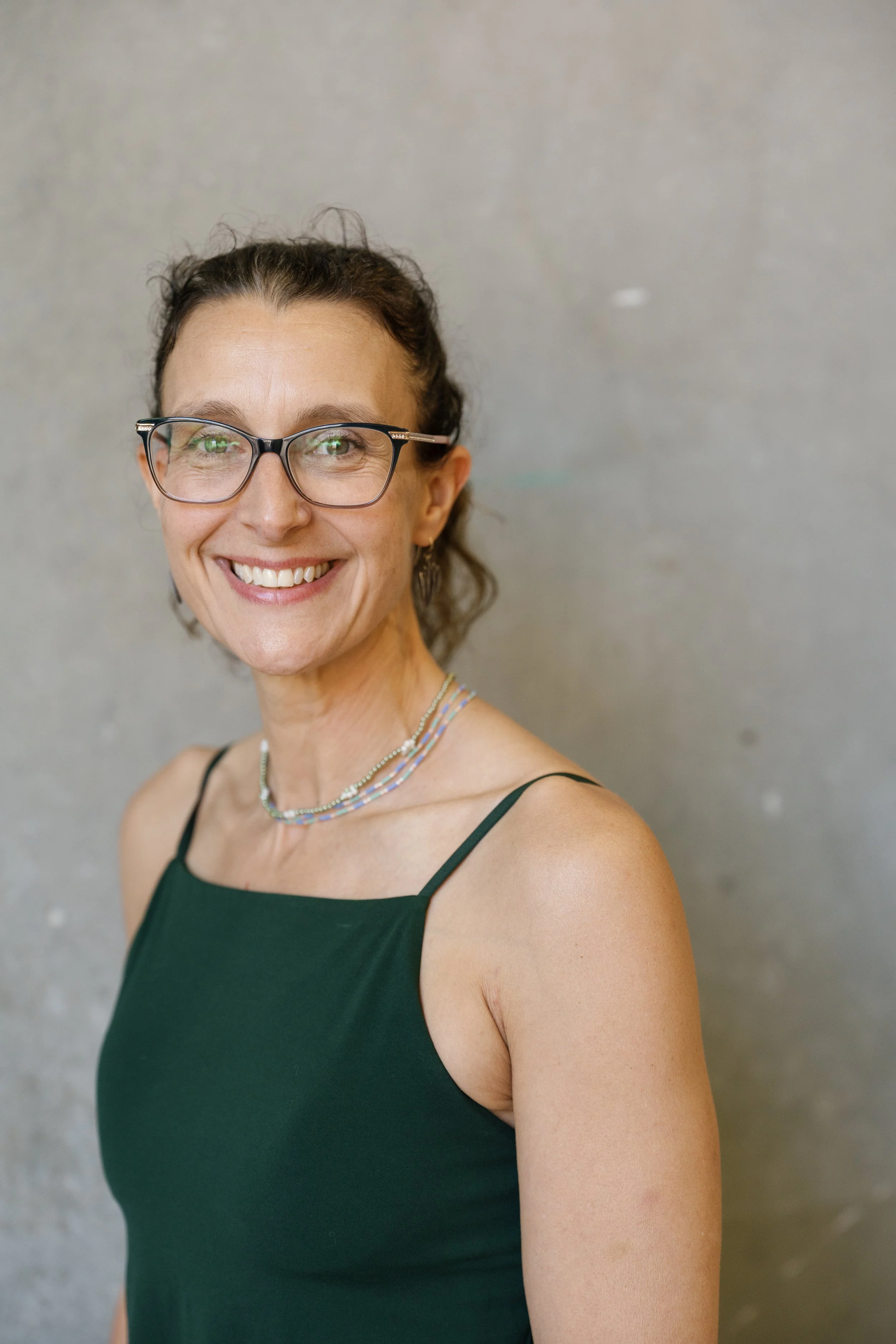 A woman smiling at the camera, wearing glasses, a dark green sleeveless top, and a beaded necklace, standing against a plain gray wall.