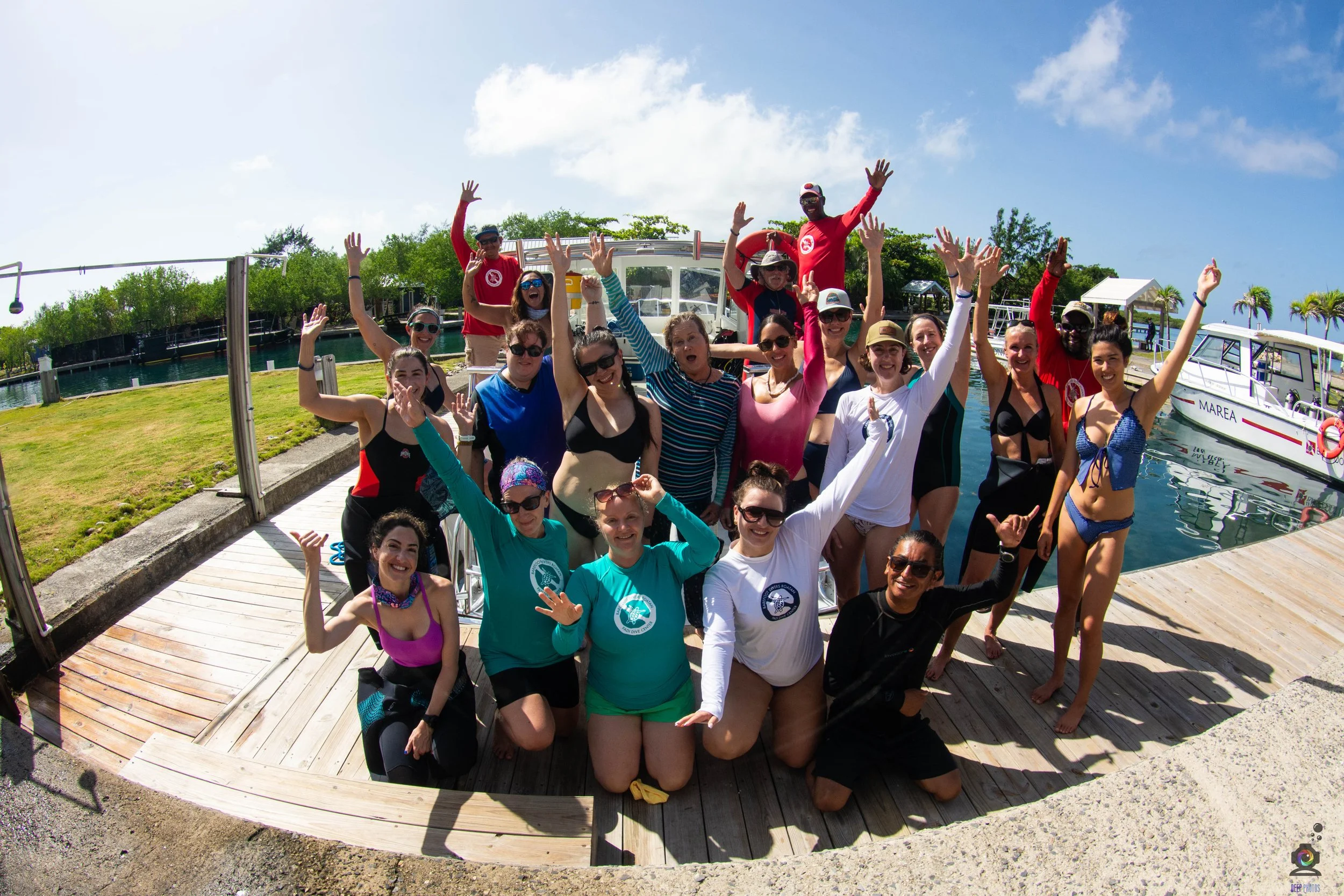 Group of people on a dock celebrating, some in swimsuits, with boats and water in the background on a sunny day.
