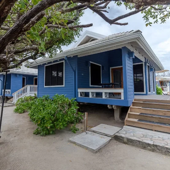 A blue beach house on stilts with a wooden staircase leading to a porch, surrounded by sand, greenery, and a large tree overhead, under a partly cloudy sky.