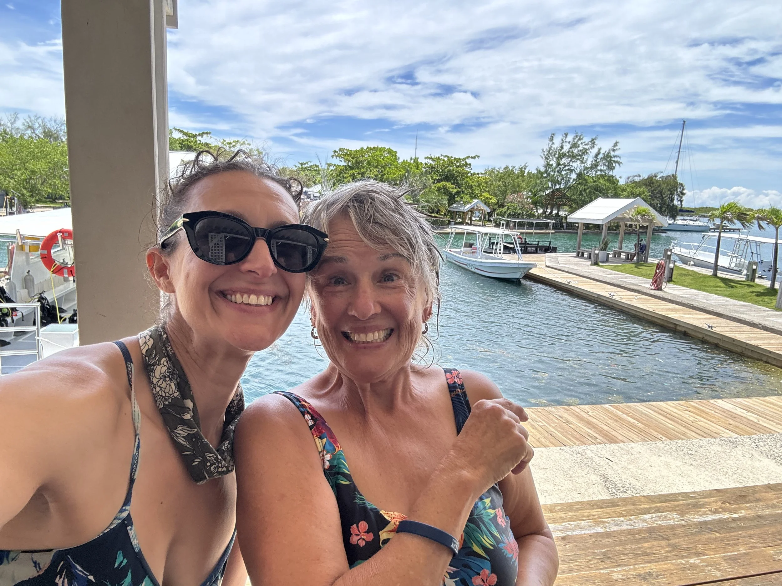Two women smiling on a dock at a marina with boats and water in the background under a partly cloudy sky.