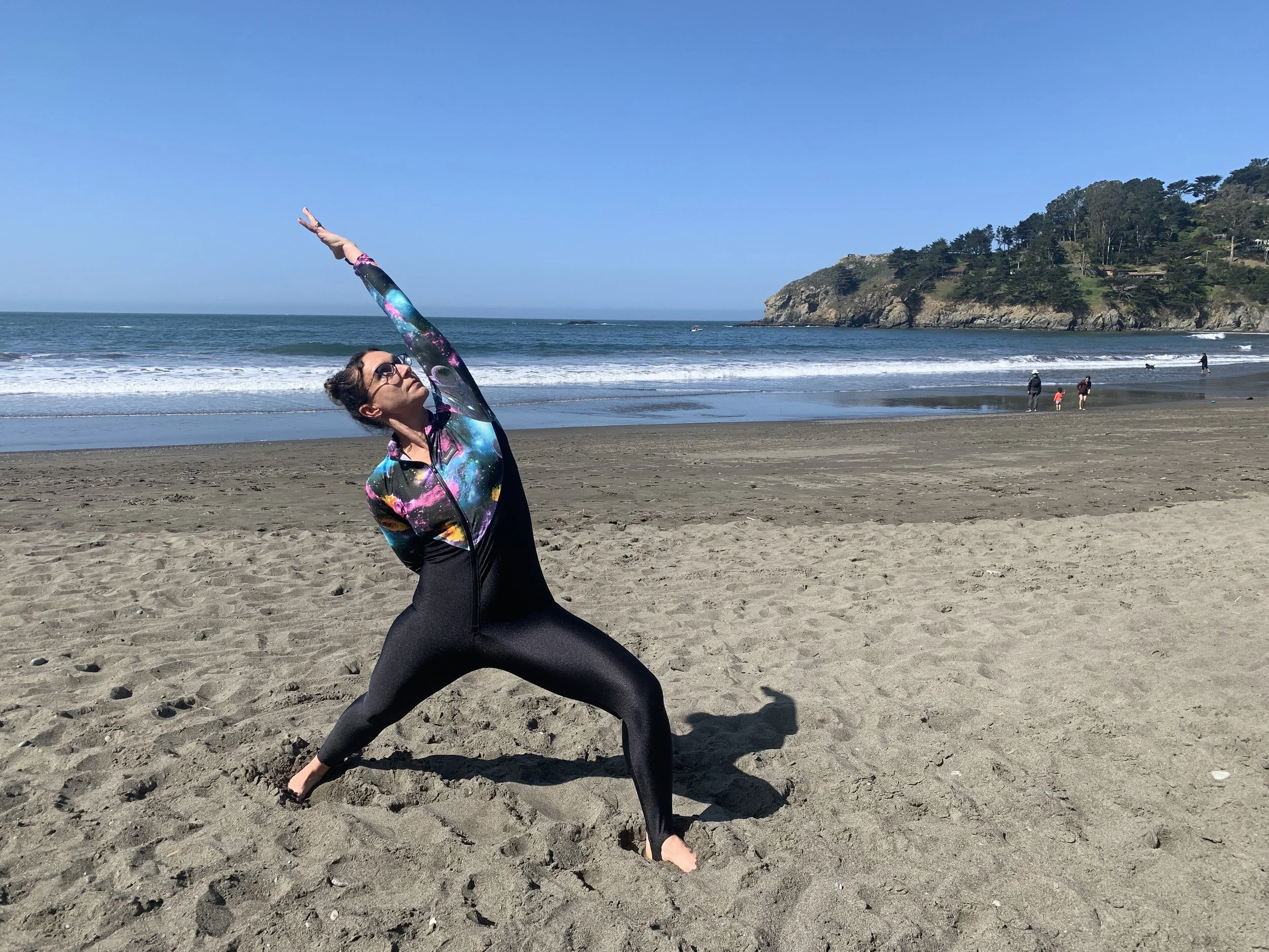Woman practicing yoga on the beach, stretching with ocean and cliffs in the background.