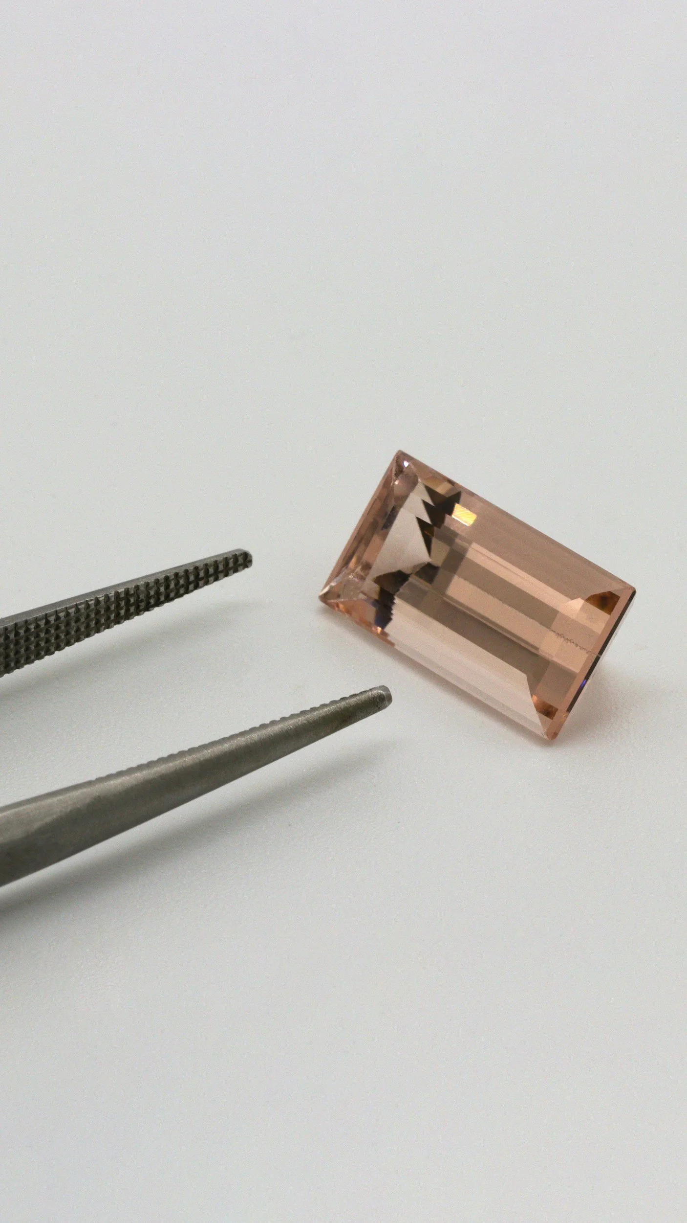 A pink rectangular gemstone held by two metal tweezers on a light surface.