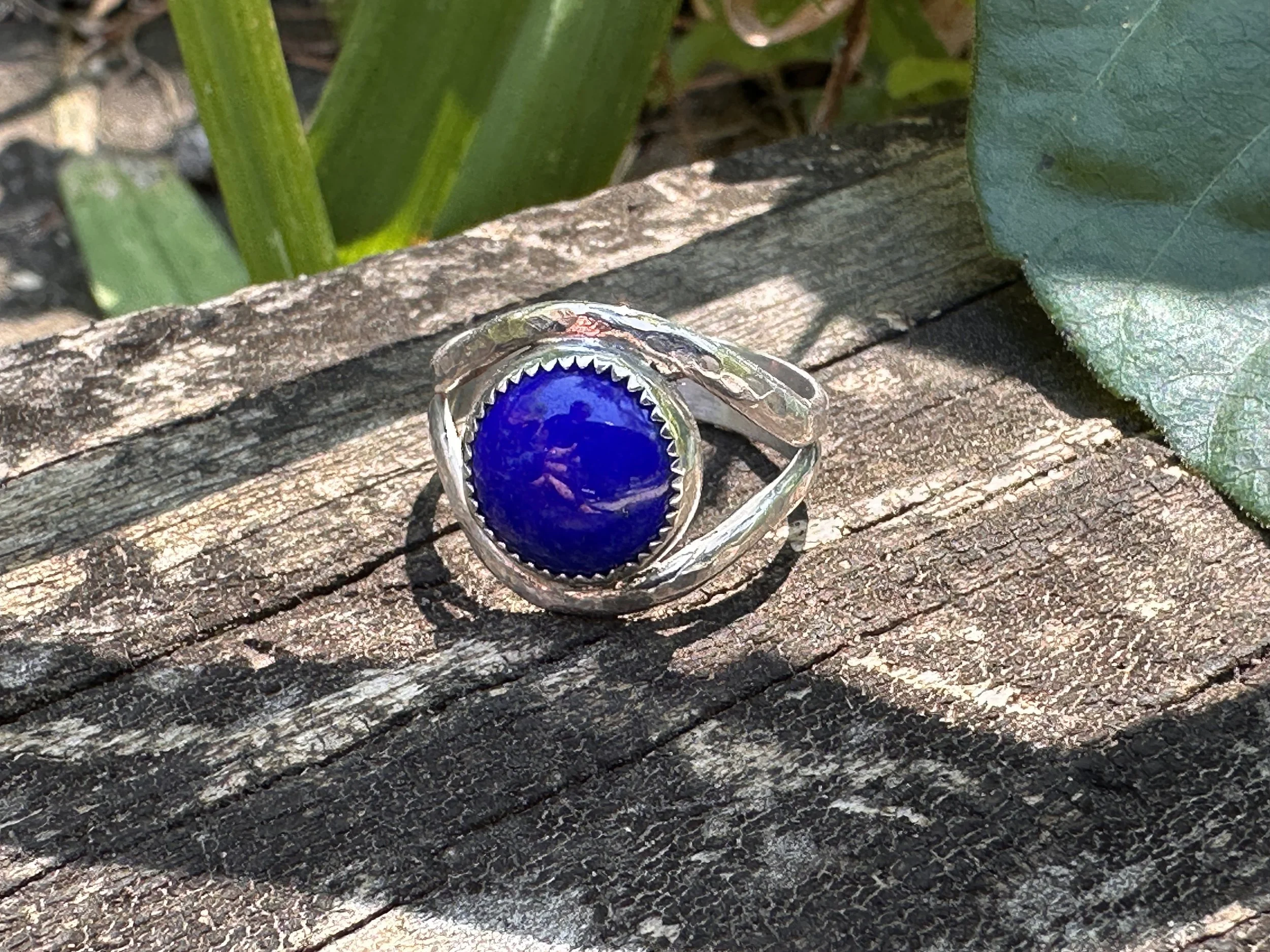 Handcrafted lapis lazuli ring with a deep blue stone in a serrated bezel on a hammered sterling silver split band, resting on weathered wood.