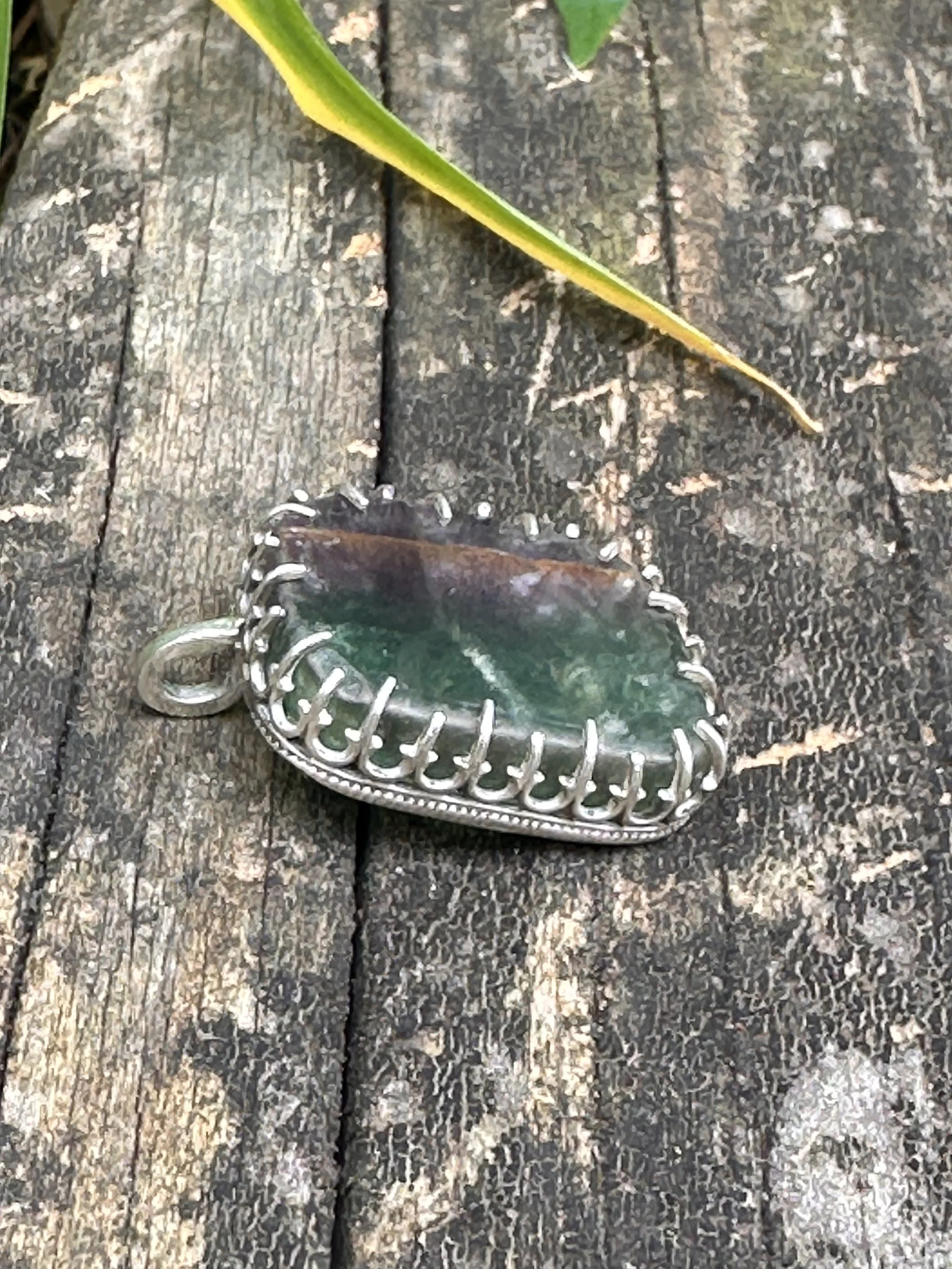 Close-up of a glass pendant with green and brown colors, lying on a weathered wooden surface, with a yellow and green leaf partially visible above it.
